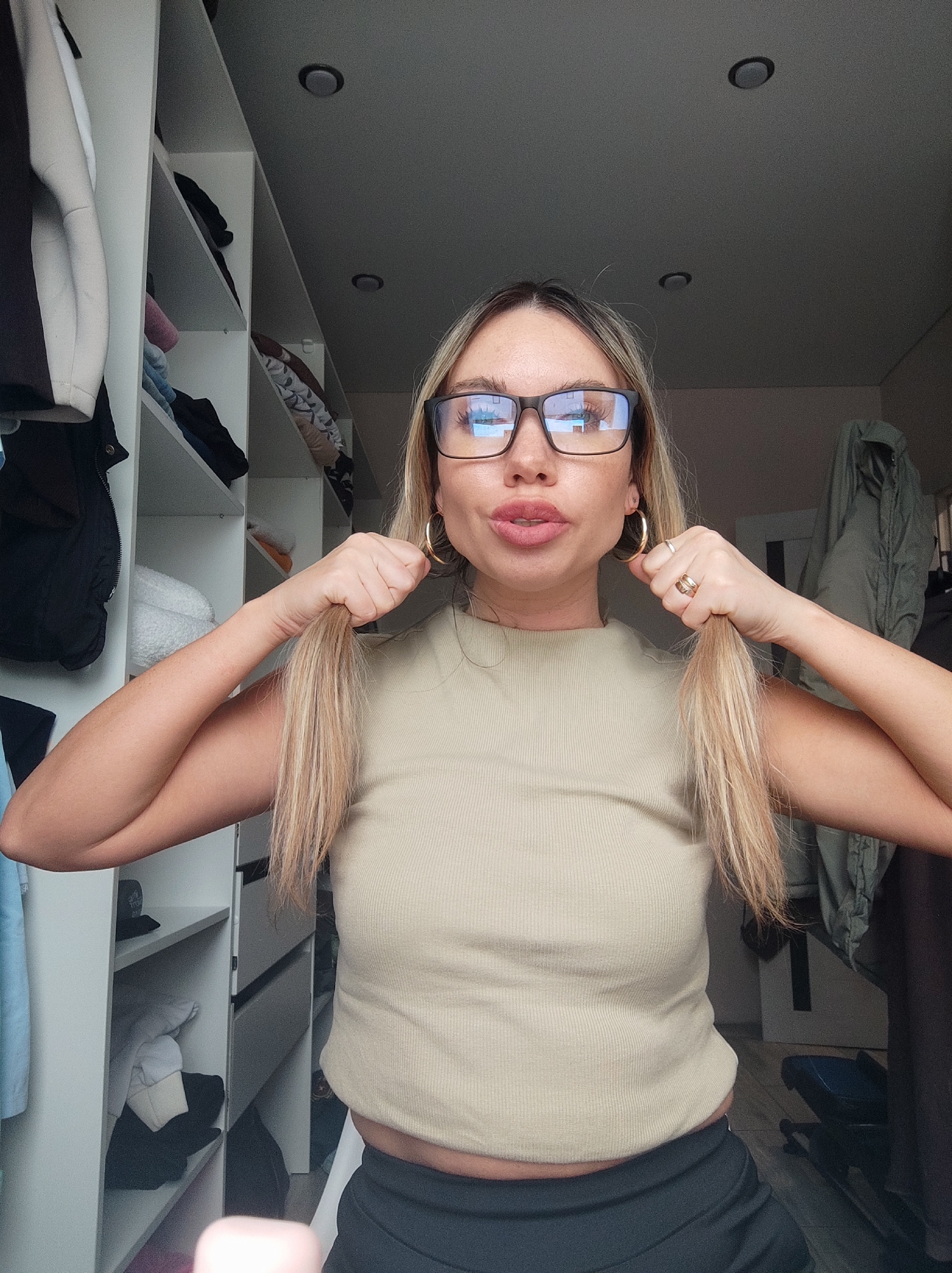 A woman is in what appears to be a walk-in closet, based on the white shelves filled with clothes visible to the left. She is wearing glasses, hoop earrings, and a tan ribbed top with black pants. She is holding the ends of her blonde hair out to either side. The lighting is dim in the upper part of the closet. The location is unknown and cannot be determined to be Votkinsk, Russia.