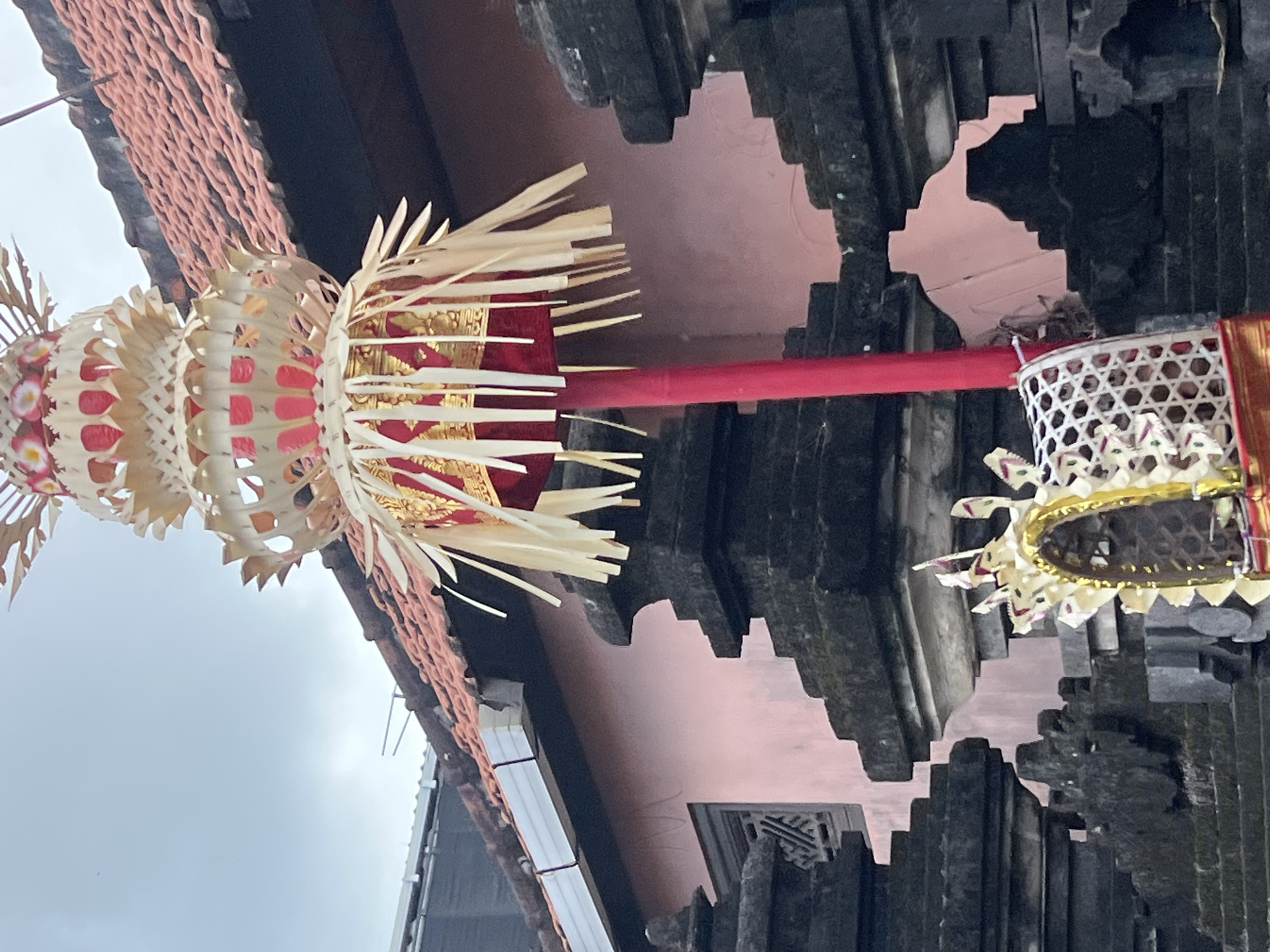 The image depicts a close-up view of traditional Balinese offerings, likely at a temple or spiritual site in Kuta, Indonesia. In the foreground, a tall, red pole supports an elaborate offering made of woven palm leaves and red fabric, adorned with gold accents. This offering is topped with multiple tiers of intricately shaped palm leaf decorations. Below this, to the right and slightly lower, is another smaller offering in a woven basket-like structure, also decorated with palm leaves and gold trim.
The background features dark, stone architectural elements that appear to be part of a temple wall or structure, with carved details. Behind the stone work, a pink-colored wall of a building is visible, topped with a traditional terracotta-tiled roof. The sky is overcast and appears to be a light grey. The scene is devoid of people. The overall impression is one of cultural and religious significance within a Balinese setting.