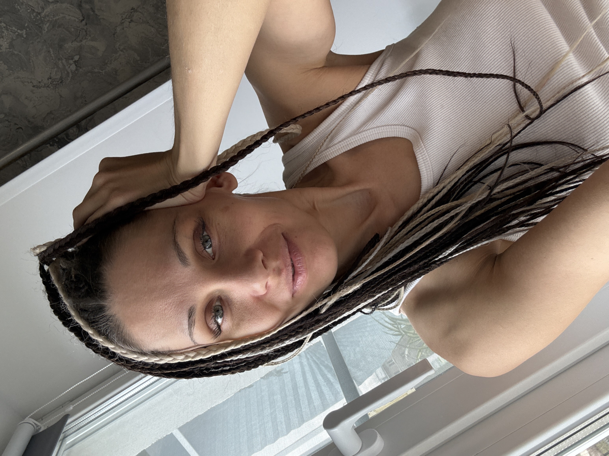 A woman is looking at the camera. She has braided hair that is brown, black, and blonde. She is wearing a white tank top and is standing in front of a window. The location is an indoor residential space, Unknown, Unknown.