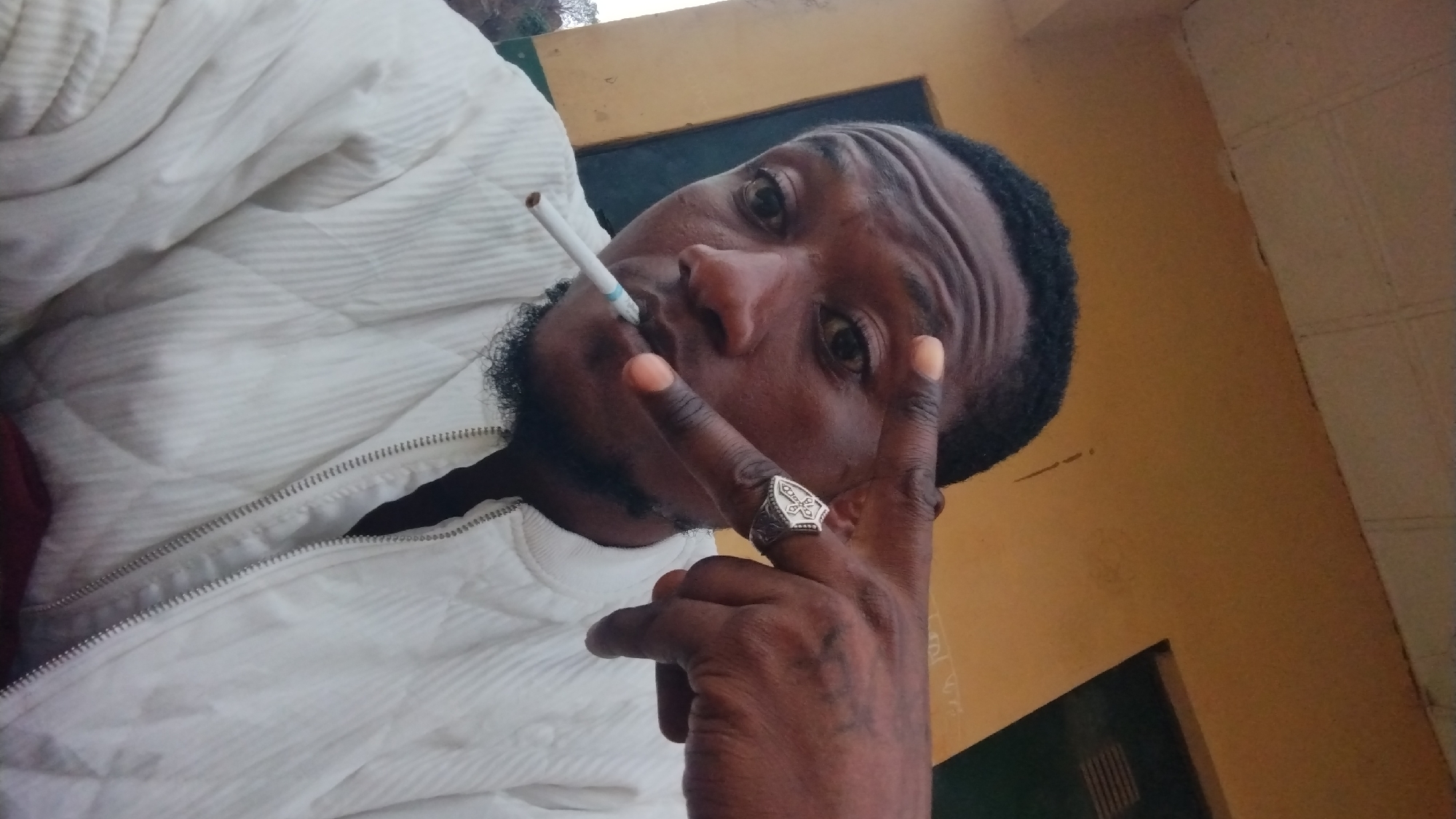 A selfie of a dark-skinned man with dark hair, a short beard, and a cigarette in his mouth. He is wearing a white quilted jacket and a silver ring on his finger, making a peace sign with his hand. The location appears to be indoors, with a wall painted yellow in the background. The location is assumed to be Jos, Nigeria.