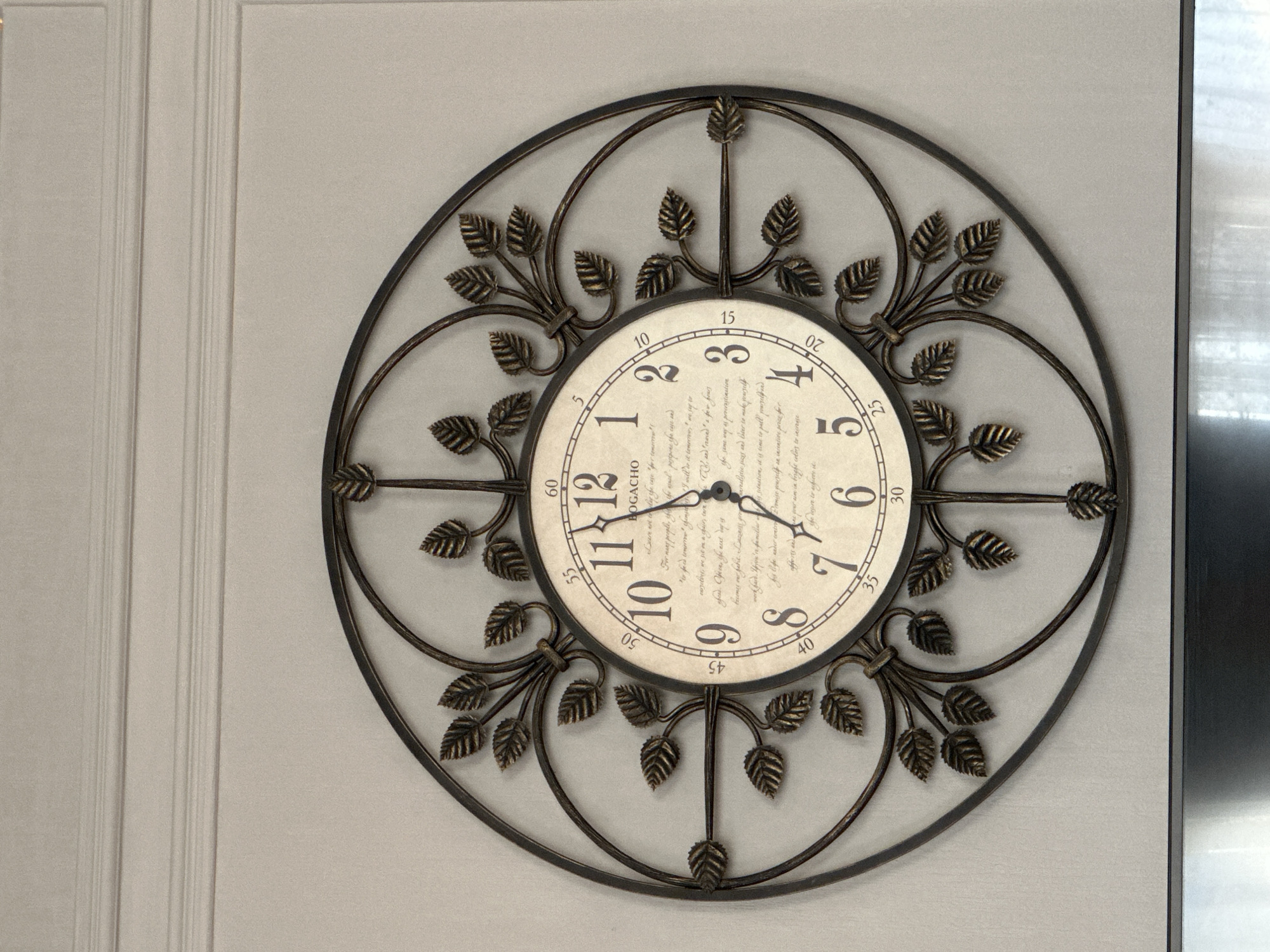 This is a close-up, eye-level, medium shot of a decorative wall clock with an ornate metal frame. The clock face is cream-colored with Roman numerals and Arabic numerals marking the hours. The brand name "OGACHO" is visible below the 12 o'clock position. The clock hands are black and appear to be at approximately 10:58. The frame is made of dark metal and is designed to resemble swirling vines with leaves. The clock is mounted on a light gray wall with subtle horizontal molding above it. The background suggests an indoor environment, possibly a room in a house or a public space. There are no people or animals visible in the image. The lighting is even, and there are no strong shadows. The city of Izobilnyy, Russia, is not visible or suggested by any elements in the image.