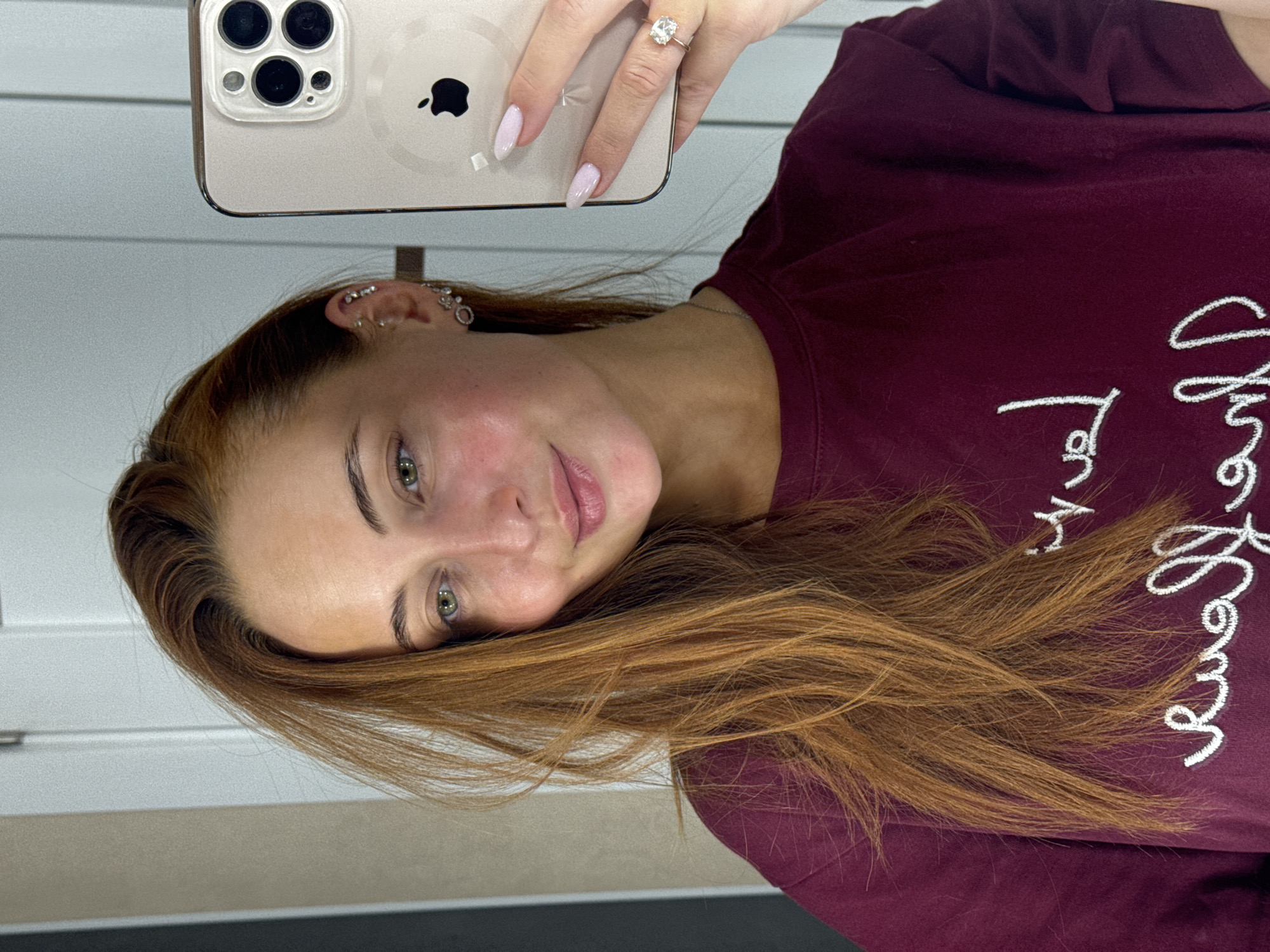 A woman with light skin, green eyes, and reddish-brown hair is holding an iPhone, taking a selfie. She's wearing a maroon sweatshirt with white embroidered writing on it. The phone case is light gold in color. She has multiple ear piercings and a diamond ring on her finger. The background appears to be an interior space with light-colored walls. There is no verifiable evidence to indicate the location is Chaikovsky, Russia.