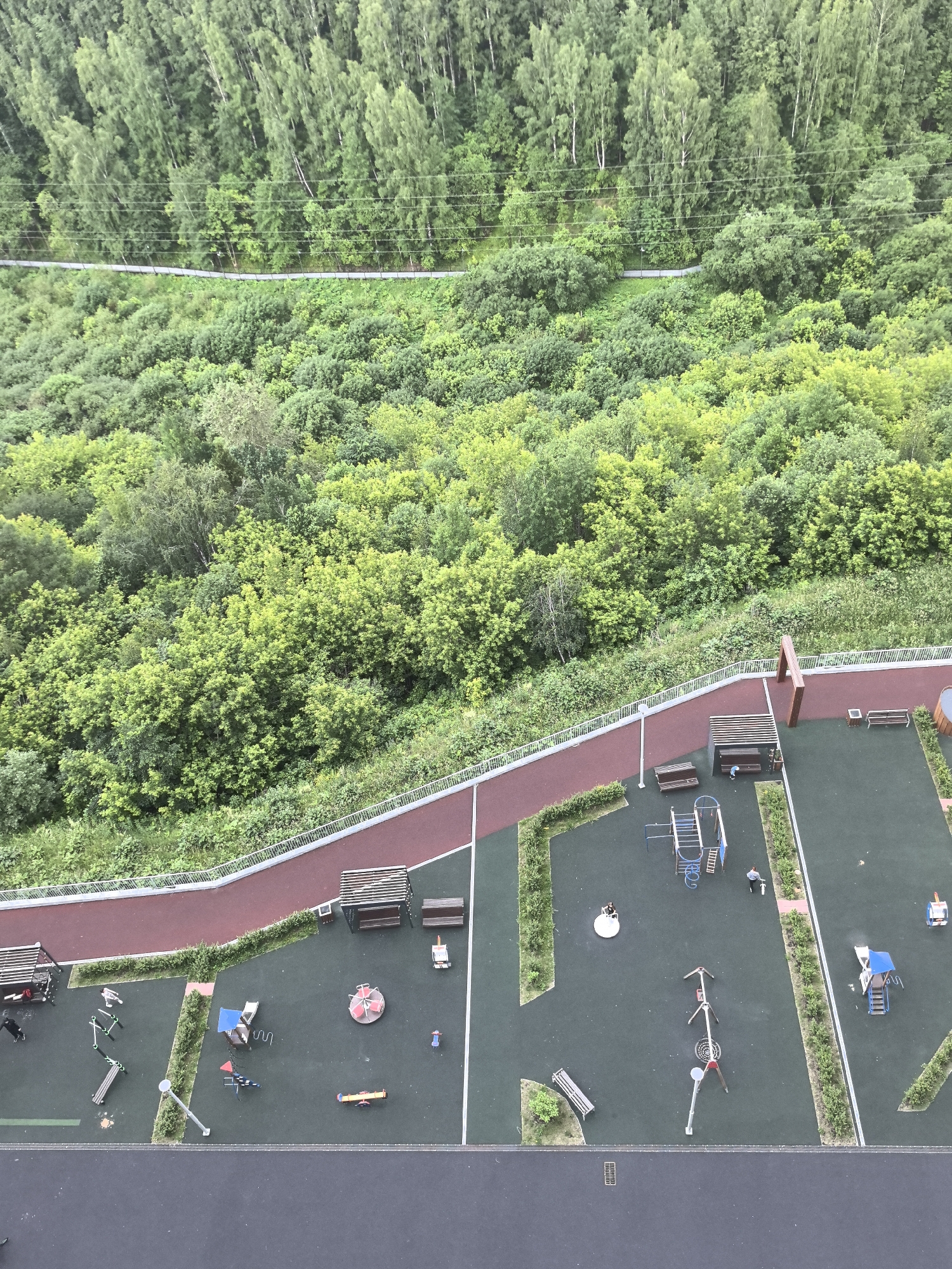 An aerial view captures a modern playground and a forested hillside in the city of Perm, Russia. The playground features green rubberized surfacing with various play structures, including swings, a merry-go-round, a seesaw, and climbing frames. Several children and adults are present, some interacting with the equipment, while others are observing or walking. A reddish-brown walking path with a white railing borders the playground. Beyond the path, a dense, verdant forest covers the hillside, with mature trees and undergrowth creating a natural backdrop. Overhead power lines are visible crossing the forest canopy. The overall scene depicts a recreational area integrated with the natural environment.