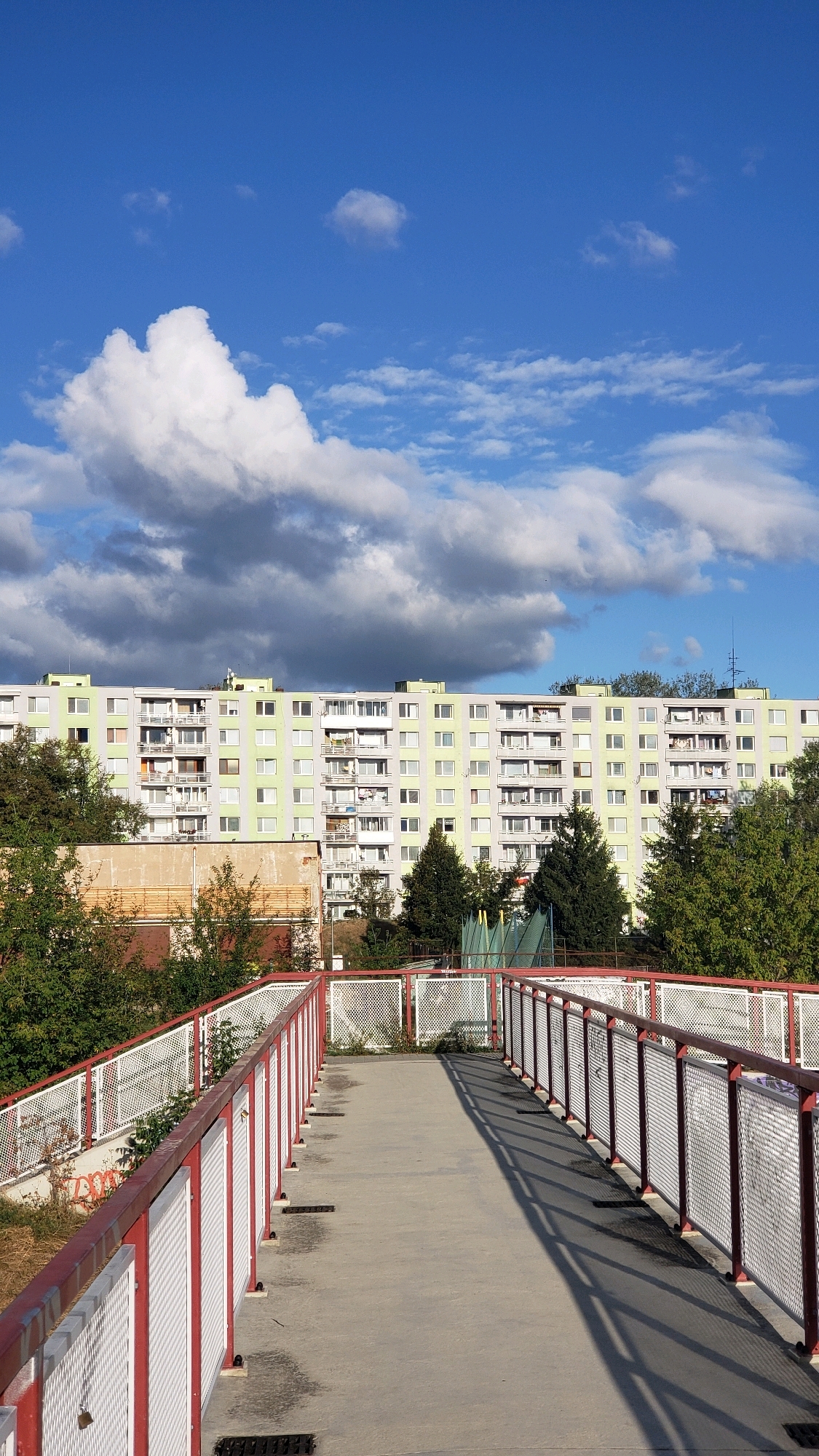 The image captures a view from a pedestrian bridge in Prešov, Slovakia, looking towards a large apartment building. The bridge, constructed with concrete and a red metal railing with white mesh infill, extends into the foreground. The apartment building, painted in a light green and white facade, features multiple stories with numerous balconies and windows, suggesting a residential block. Lush green trees and some smaller buildings are visible in front of and around the apartment building. The sky above is a vibrant blue, adorned with large, fluffy white clouds and some smaller, scattered clouds. The overall scene depicts an urban environment under a bright, partly cloudy sky.