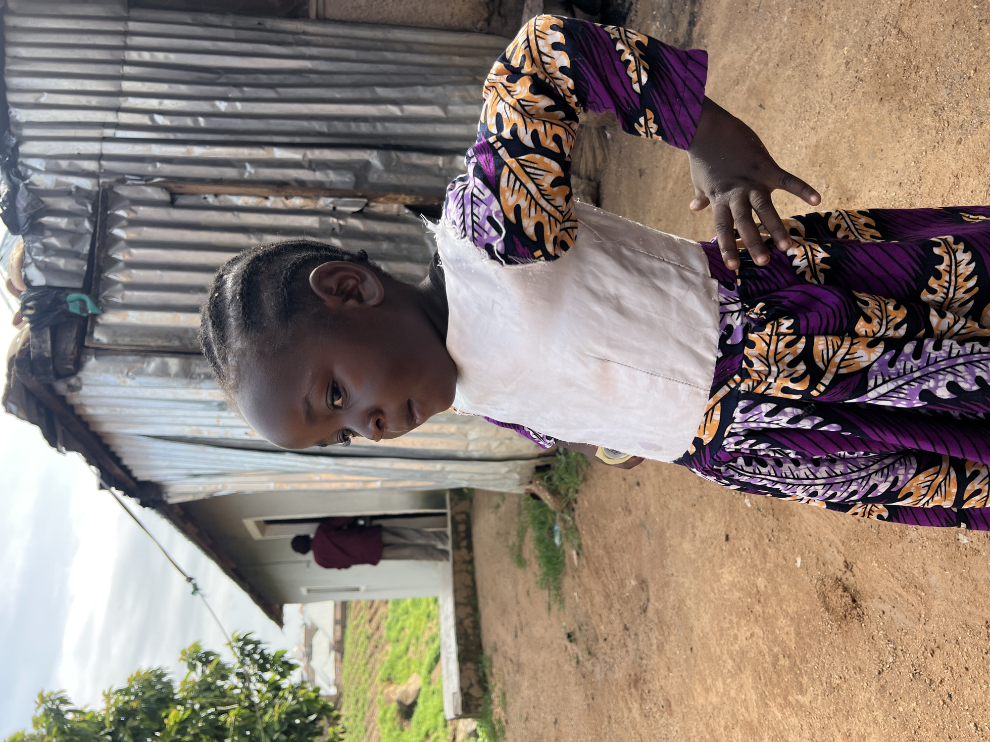 A young Black child stands facing left in the foreground, looking into the distance. The child wears a white top with colorful, patterned sleeves featuring purple, orange, and black leaf designs. The child's skirt is also patterned with the same design. The child's left hand is on their hip, with fingers slightly spread.
Behind the child, a building with a corrugated metal roof and sides is visible. To the left of the metal building, another building with light-colored walls and a lighter roof can be seen. A person in a maroon shirt and tan pants is standing in the doorway of the lighter-colored building, facing away from the camera. The ground is a mix of reddish-brown dirt and sparse green grass. In the background, rolling green hills with scattered buildings can be observed. The sky is overcast with a few visible clouds. The scene appears to be an outdoor residential area in Jos, Nigeria, likely a settlement or informal housing area given the building materials.