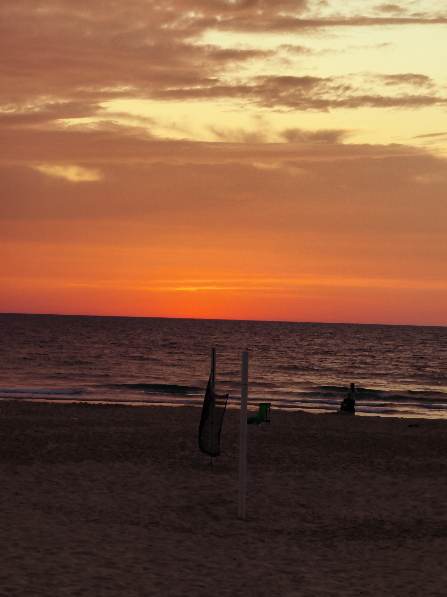 This media file depicts a serene sunset over a beach, likely in Ashkelon, Israel. The sky is dominated by a breathtaking display of warm colors, with a vivid gradient of fiery orange and deep red near the horizon where the sun is setting, transitioning into softer yellows and light oranges higher up, interspersed with scattered, elongated clouds.
The sea stretches across the mid-ground, a darker expanse of water with gentle waves breaking near the shore, reflecting subtle hints of the sky's colors. In the foreground, the sandy beach appears dark, almost silhouetted, consistent with the low light conditions of dusk.
Key objects include a white pole from a volleyball net, with a dark net partially visible, situated on the left side of the beach. A small, bright green chair is also visible behind the net pole. On the right, a solitary figure is seated on the sand at the water's edge, presented as a dark silhouette, seemingly observing the tranquil scene or meditating by the sea.
The image captures the peaceful transition from day to night, characterized by the striking natural light and the calm ambiance of the beach. There is no visible text within the image.