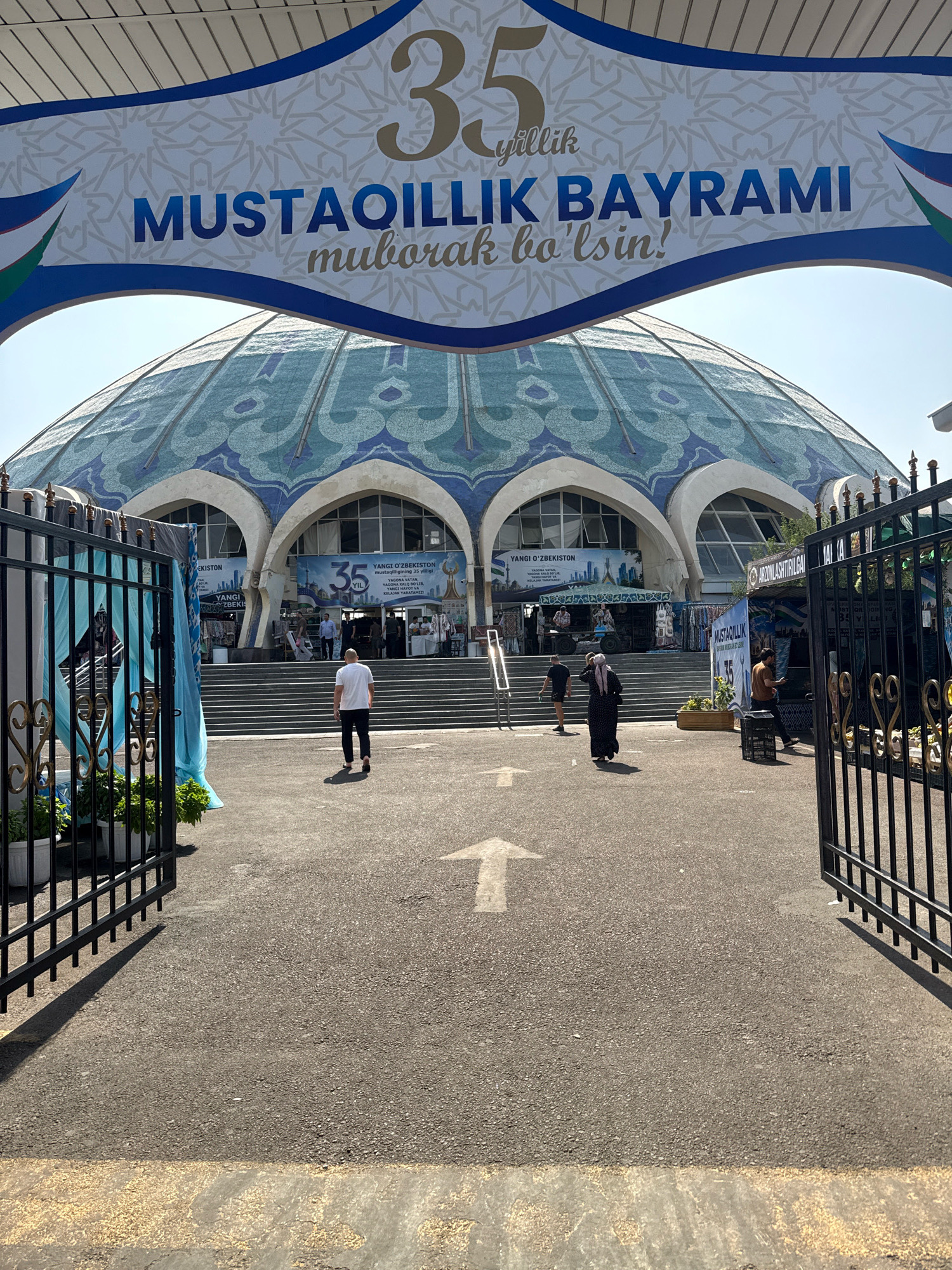 The image shows the entrance to the Chorsu Bazaar in Tashkent, Uzbekistan. A large, decorative banner hangs overhead, featuring the number "35" and text that reads "MUSTAQILLIK BAYRAMI muborak bo'lsin!". This translates to "Happy Independence Day!". The banner is draped over a large, domed building with intricate blue and green mosaic tilework, characteristic of traditional Uzbek architecture.
In the foreground, a black metal gate is open, leading onto an asphalt pathway marked with a white arrow pointing forward. Several people are visible: a man in a white t-shirt and black pants walks away from the camera towards the bazaar entrance. Another person, wearing a dark dress and headscarf, walks in the same direction. Further back, several individuals can be seen near the entrance steps and inside the market stalls. Various banners and signage, including one with "35 YIL" (35 years) and "YANGI O'ZBEKISTON" (New Uzbekistan), are displayed around the entrance, indicating a celebration. The overall environment suggests a bustling market on a sunny day, with the architecture hinting at a culturally significant location.