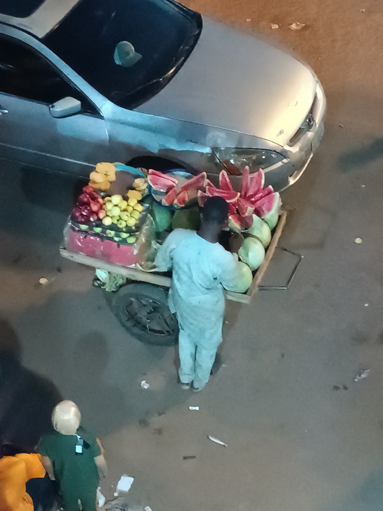 The scene depicts an overhead shot of a street in Bukuru, Nigeria. The street surface is gray, and scattered debris litters the ground. A silver-colored car is parked along the edge of the street. A man dressed in light-colored clothing stands behind a pushcart piled high with various fruits including watermelons. There are two other individuals visible in the bottom left corner of the frame. One is wearing a green shirt and the other is partially obscured but appears to be wearing an orange shirt. No buildings or other infrastructure are clearly visible in the background.