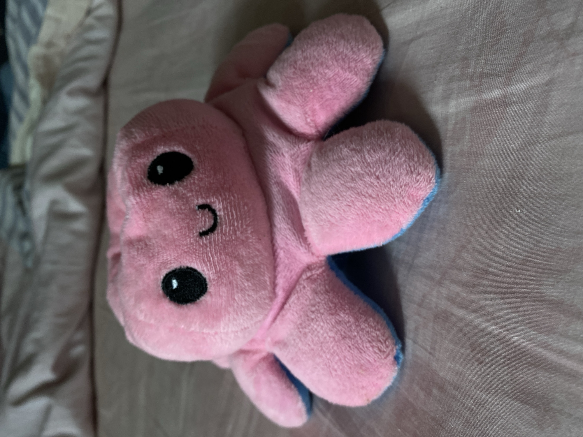 A pink plush octopus toy with blue accents on its tentacles sits on a pinkish-gray textured surface, possibly a bedspread. The octopus has two black button eyes and a small, curved black smile. The toy is centrally framed, with its tentacles splayed outwards. The background is blurred, showing folds and textures of fabric. The lighting suggests an indoor setting.
