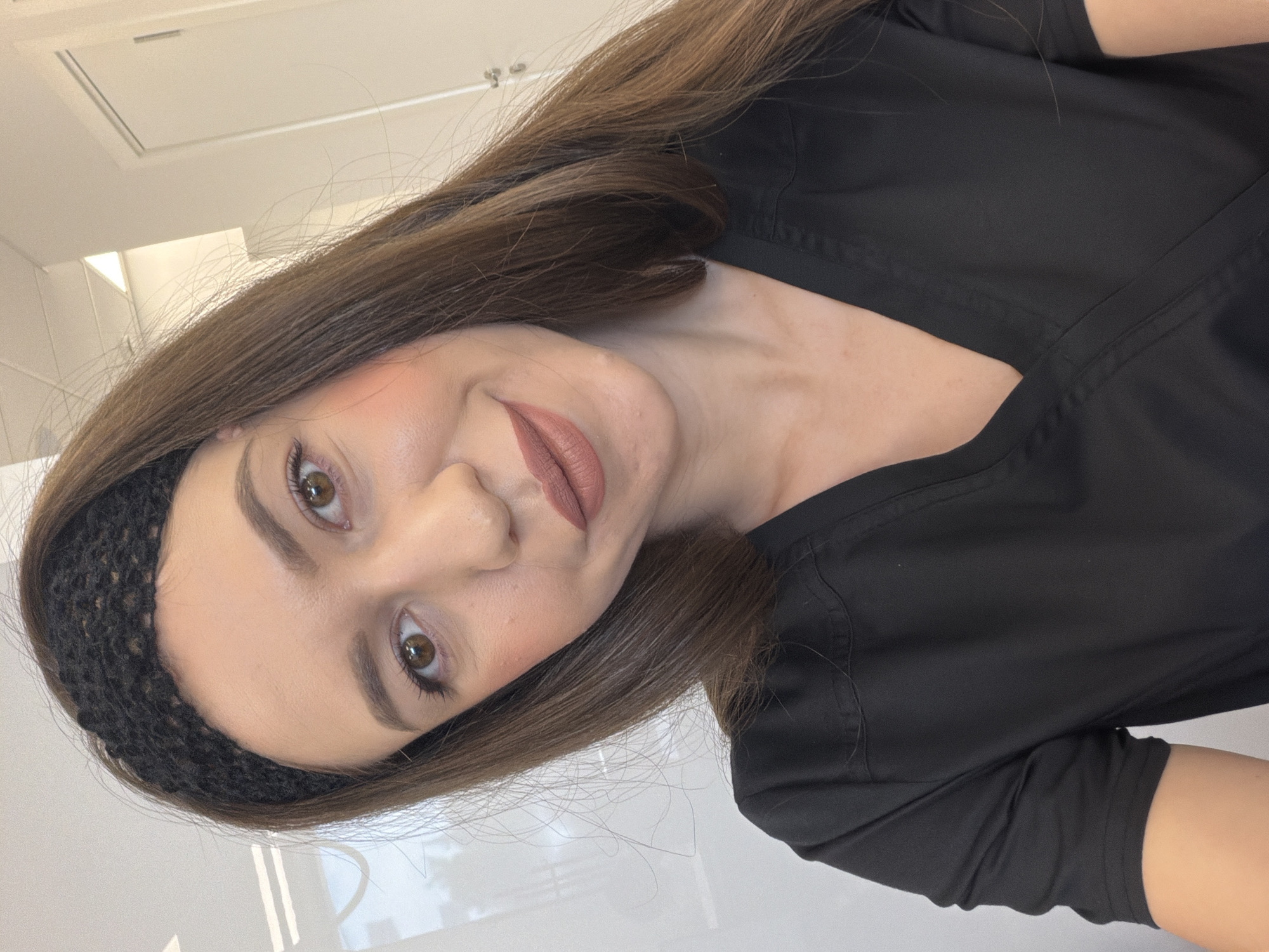 A woman with long, brown hair and a black knitted headband poses for a selfie indoors. She is wearing a black scrub top. Her makeup includes blush on her cheeks and a matte brown lipstick. The background appears to be a well-lit room with white walls and doors. The image was taken in Sofia, Bulgaria.