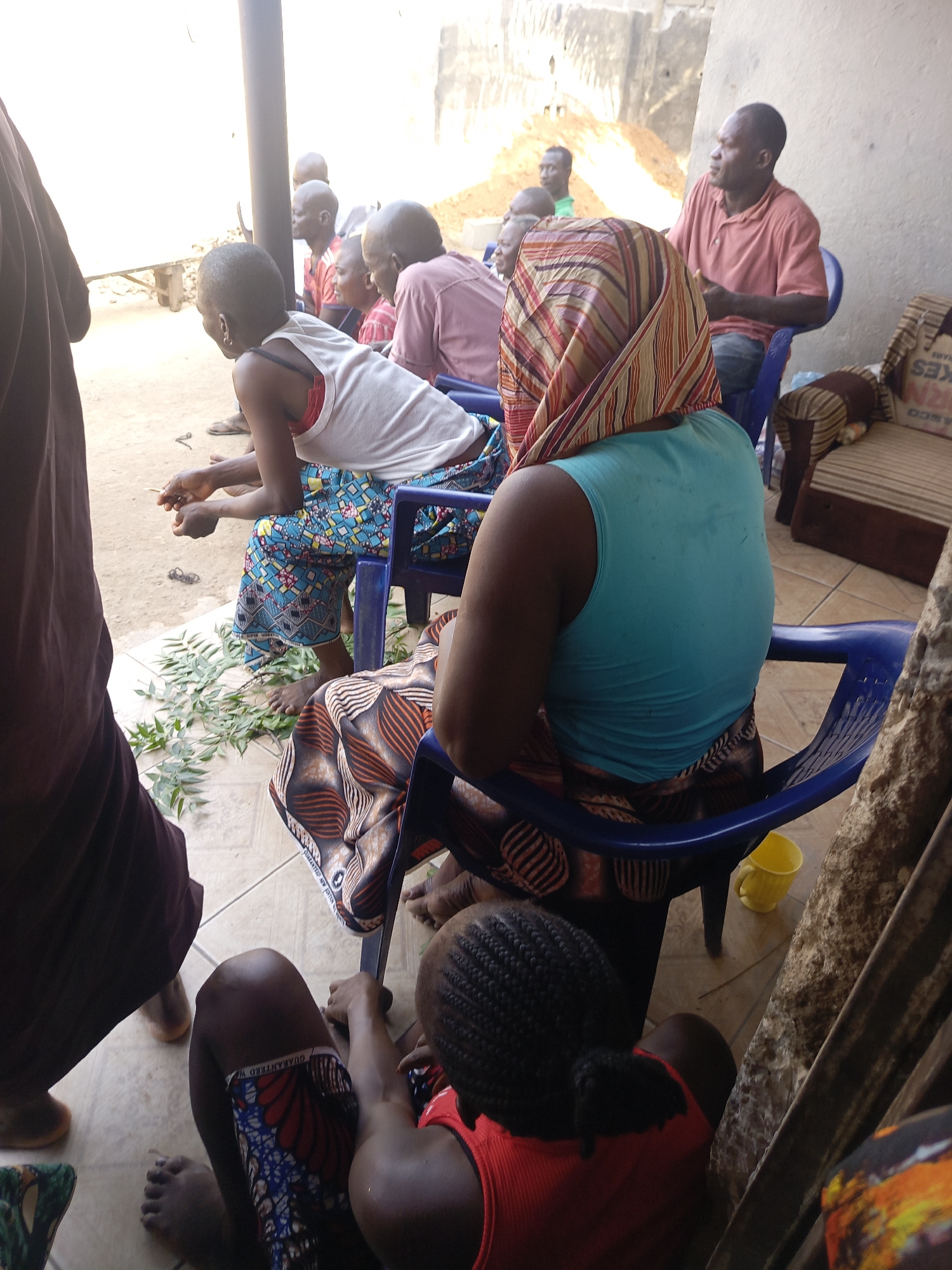 The image depicts a group of people gathered in what appears to be an outdoor setting in Akwanga, Nigeria. Several individuals are seated in blue plastic chairs, while one person is sitting on the floor. A woman in a turquoise tank top and patterned headscarf sits with her back to the camera. A young person wearing a red shirt sits on the ground beside her. Scattered green leaves are visible on the tiled floor. The environment appears to be a partially covered courtyard or veranda. Additional details include a sofa with striped cushions and an old box in the background, along with glimpses of the exterior wall. The scene conveys an informal gathering, possibly a social or familial event.
