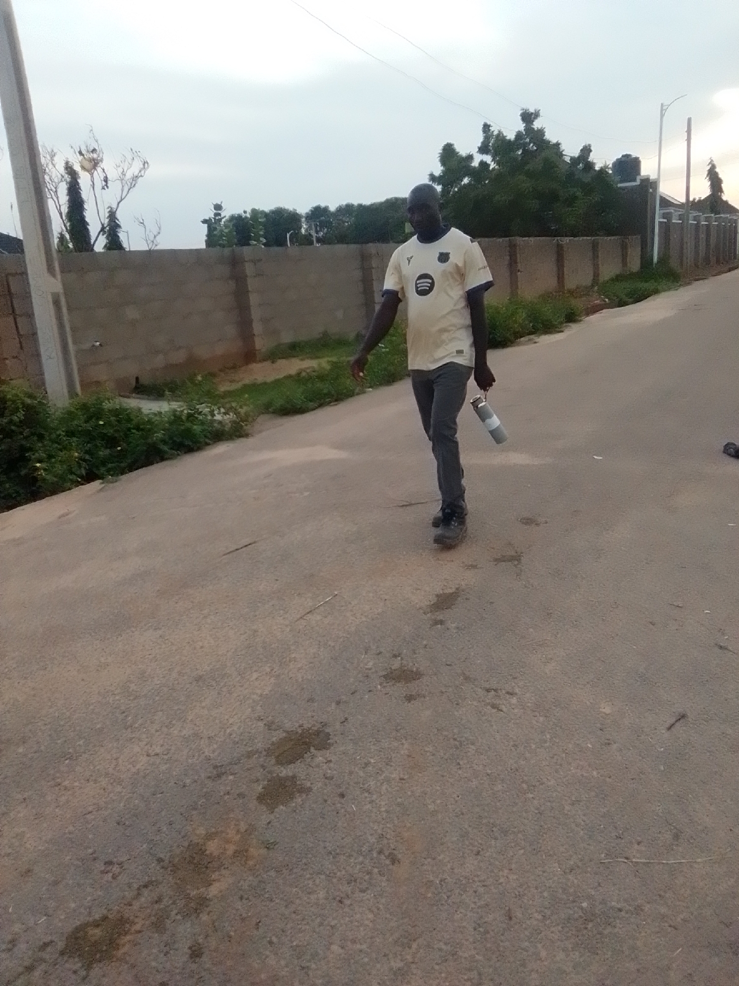 A lone Black male walks along a paved road in Liji Hill, Nigeria. He is wearing a light-colored t-shirt with a logo on the chest and dark pants, and carrying a silver thermos in his right hand. The road is flanked by a low concrete wall and some vegetation. Trees and buildings are visible in the background under an overcast sky.