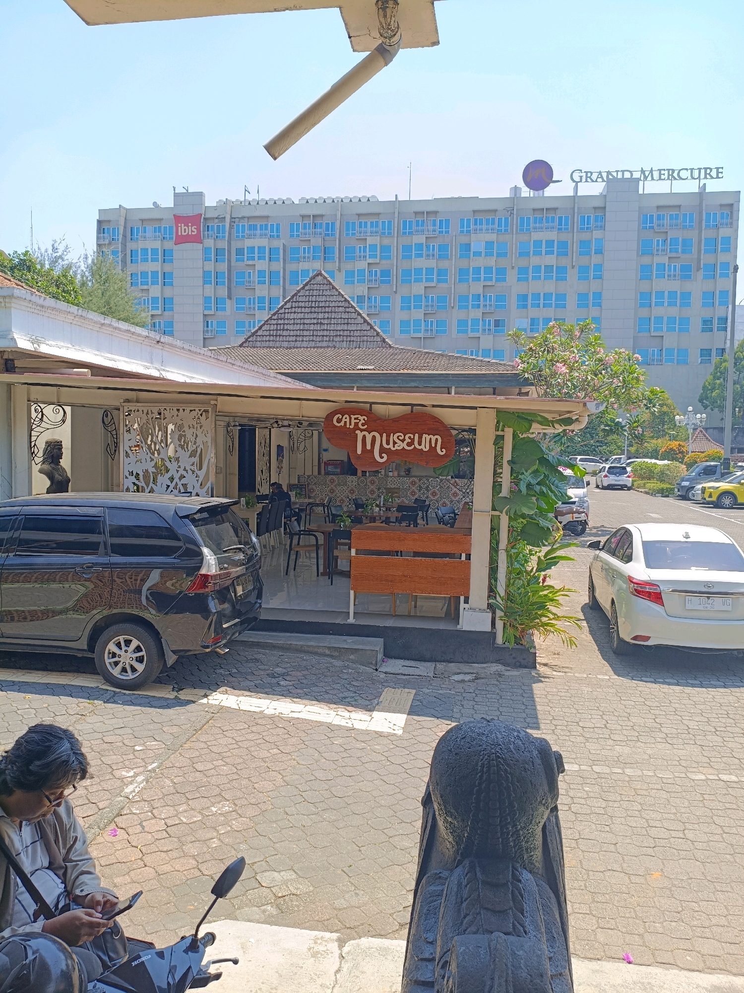 This image captures a street view in Depok, Indonesia, on a sunny day. In the foreground, a person is seated on a motorcycle, looking at their smartphone. The motorcycle is black with "HONDA" visible on its body. The ground is paved with cobblestones.
Behind the person and motorcycle, a black SUV is parked on the street. Further back, a white sedan is parked in a lot. A sign that reads "CAFE MUSEUM" is prominently displayed on a small building. The building has white lattice work on its facade and a tiled roof.
In the background, a large hotel complex is visible. One part of the hotel displays the "ibis" logo in red, and another part shows the "GRAND MERCURE" signage. The hotel has a modern facade with numerous blue-tinted windows. Trees and foliage are scattered throughout the scene, indicating an urban setting with green spaces. The overall impression is of a vibrant, well-lit urban environment.
