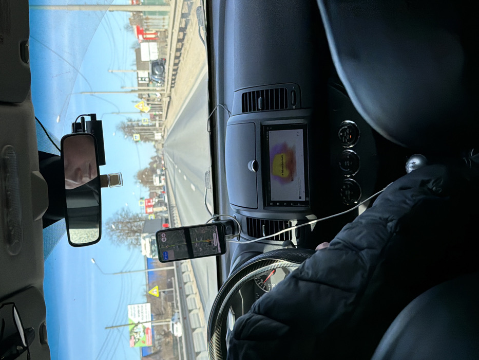 The image depicts the interior view from the passenger seat of a vehicle traveling northbound on a multi-lane highway in Zavidovo, Russia, under clear daylight conditions.
**Vehicle Interior:** The driver, whose face is partially reflected in the rearview mirror, has their left arm, clad in a dark quilted jacket, positioned on the steering wheel, which features a carbon fiber-textured cover. A smartphone, displaying a map navigation application with a green highlighted route, is mounted on the dashboard and connected via a white charging cable. Below it, the vehicle's infotainment system shows a colorful graphic with text partially legible as "Move this soul." A manual gear shift lever is visible in the lower center console. A dashcam is mounted near the rearview mirror.
**Exterior Environment:** The vehicle is on a paved road with at least two lanes in each direction, clearly marked by solid and broken white lines. A metal guardrail runs along the left side of the road. On the right shoulder, the ground is dry, with sparse brown vegetation and dirt. Utility poles with power lines line both sides of the road.
**Roadside Features:** On the left, a large billboard with Cyrillic text is visible, along with a white semi-truck in the distant opposing lane. On the right, a yellow triangular warning sign, green rectangular road signs, and additional utility poles are present. Further along the right side of the road, several low-rise commercial buildings with red roofs and white facades are visible. A dark-colored off-road vehicle is parked on the unpaved shoulder. The sky is clear blue, and trees in the background appear bare, suggesting late winter or early spring.