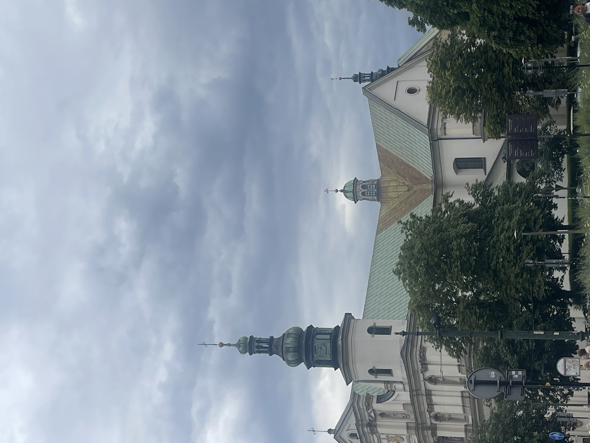 A low-angle, medium shot captures a historic building with multiple spires and green, sloped roofs against a cloudy, overcast sky in Krakow, Poland. The architecture appears to be ecclesiastical, possibly a church or monastery. One prominent tower, painted white with green accents, rises to the left, topped with a green, tiered spire. To the right, a second, smaller green spire is visible above the roofline. Trees with lush green foliage are in the foreground and around the building. A street light pole with signs is positioned in the lower-left quadrant of the frame. A few people are faintly visible in the lower portion of the image, in front of the building and trees. The overall impression is of a stately, historical edifice under a somber sky.