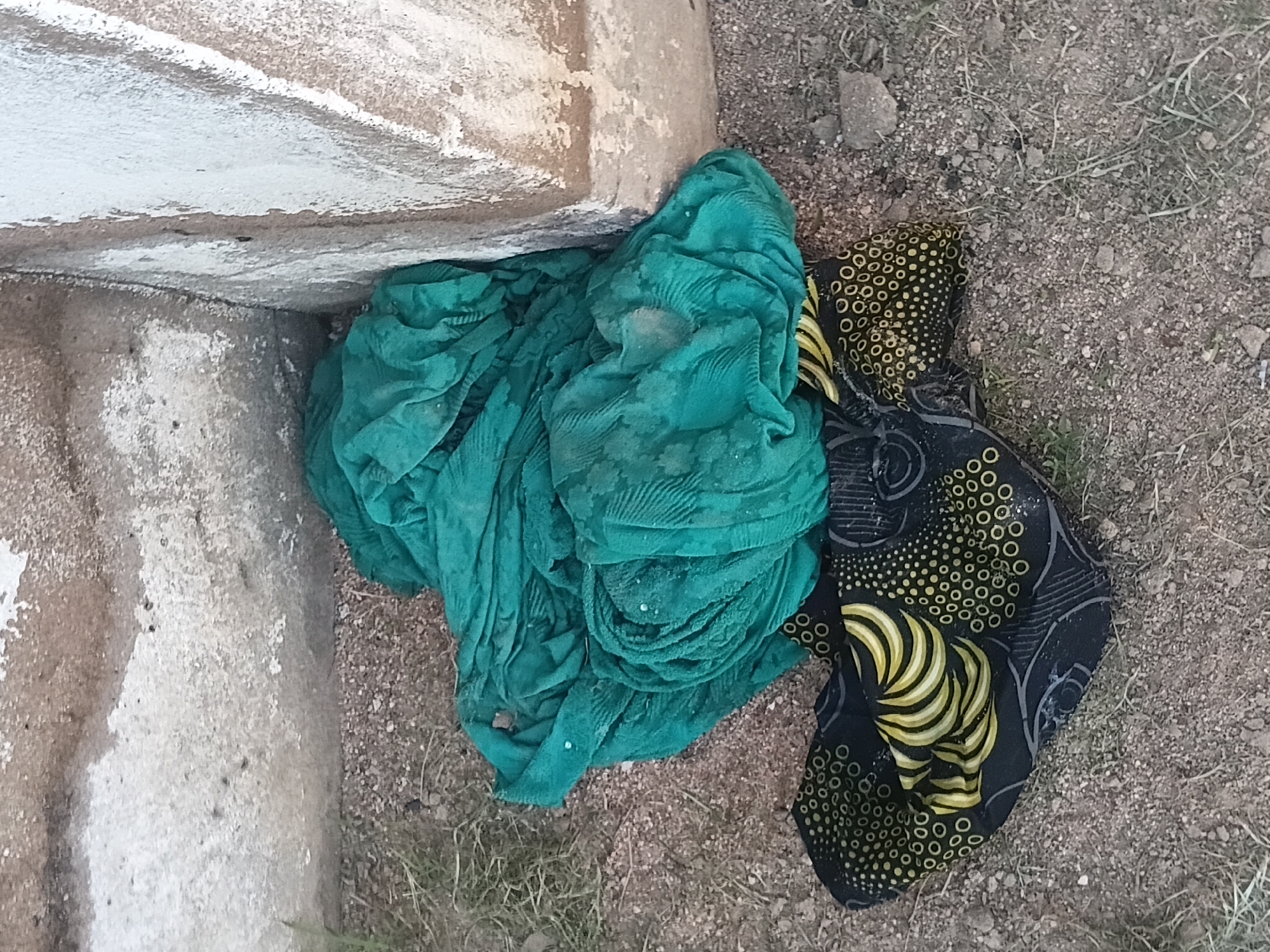 The image shows two pieces of fabric lying on the ground. The larger piece is a teal-colored, folded cloth with a textured pattern. The smaller piece, beneath the teal cloth, is black with a yellow and white circular dot pattern and wavy yellow stripes. The fabrics are situated next to a rough, concrete structure, possibly a wall or building foundation, with patches of dirt and sparse grass around it. The ground is covered in small pebbles and dirt. The setting appears to be an outdoor, possibly urban or semi-urban environment, given the presence of the concrete structure. Without further context, the specific location within Jalingo, Nigeria cannot be determined from the image alone.