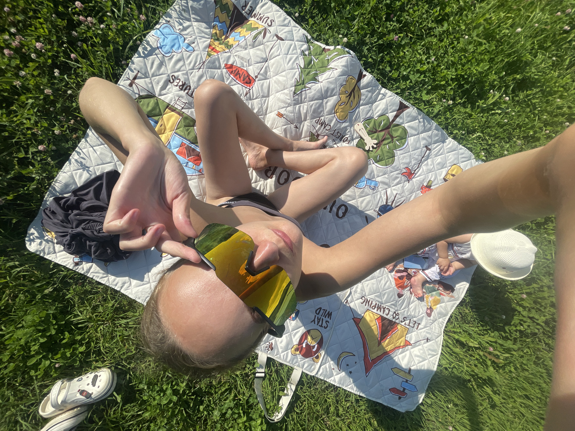 The image shows a person lying on a patterned blanket spread on a grassy area. The blanket has a "Forest Camp" theme with cartoonish illustrations of tents, trees, and other camping elements. Text on the blanket includes "SUMMER," "STAY WILD," and "LET'S GO CAMPING." The person is wearing sunglasses with mirrored yellow lenses and is positioned to take a selfie. Their legs are bent, and one arm is raised, holding the sunglasses. A dark piece of clothing is on the blanket near the person's legs, and a pair of white clogs are on the grass next to the blanket. In the background, a white straw hat is visible on the blanket, along with what appears to be another person or child lying down, wearing striped shorts and a white shirt. The scene is illuminated by bright sunlight, suggesting a clear, sunny day. The overall impression is of a leisurely outdoor activity, likely a picnic or a relaxing day in a park or backyard in Подшивалово, Russia. The mood appears relaxed and cheerful, with the person enjoying the warm weather.