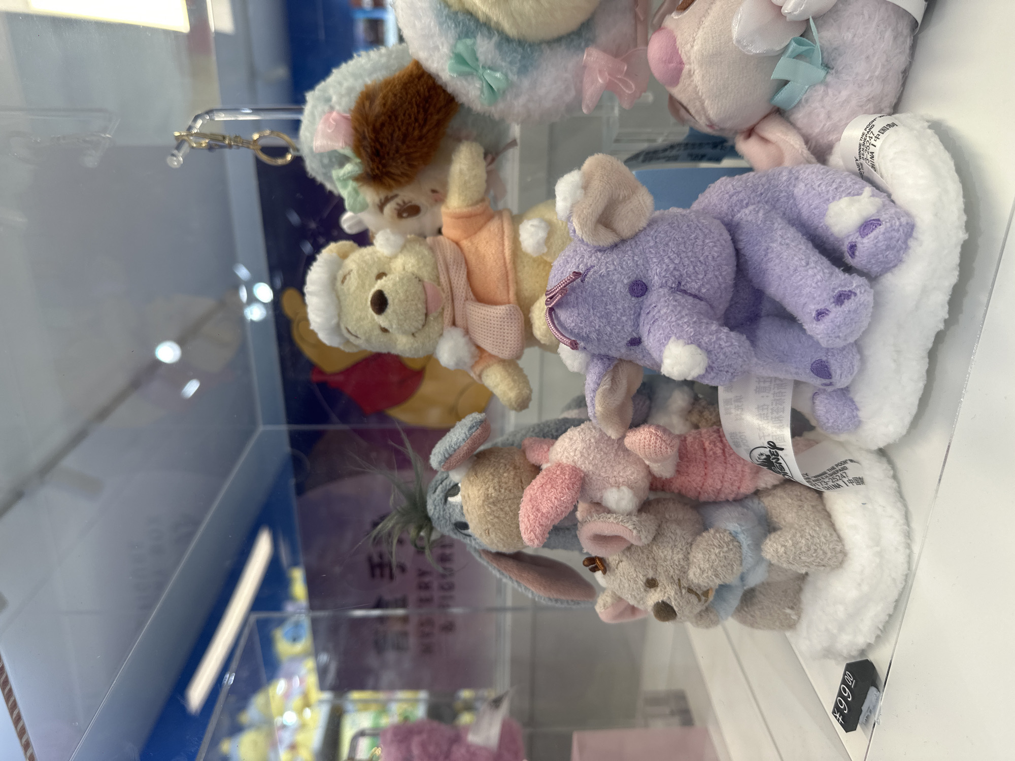 In a retail display case, likely located in Futian District, China, several plush toys are arranged on white cloud-like bases. From left to right, a grey Eeyore stuffed animal with pink inner ears is prominently displayed, holding a small pink piglet-like plush. To the right of Eeyore, a yellow Winnie the Pooh plush wearing an orange shirt and a pink scarf is visible. Partially obscuring the Winnie the Pooh plush is a light blue plush with brown fur and a pink bow. In the foreground, a purple elephant plush with pink inner ears sits on a white cloud base. To the far right, a pink plush with a light blue bow is partially visible. A price tag displaying "¥99.00" is positioned on the left side of the display. The background is blurred, showing shelves and a blue-colored wall with some text that appears to be in Asian script. The display case is made of glass, and reflections from overhead lighting are present.