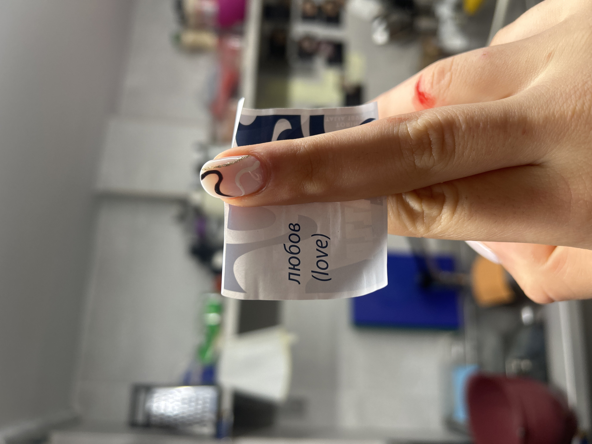 A hand holds a small piece of paper with the word "любов" (love) written in Russian and English. The hand has a beige manicure with black and white swirls. There is a small wound on one of the fingers. The background is blurry but suggests a kitchen or laboratory setting. It is difficult to determine if this image was taken in the city of Burgas, Bulgaria.