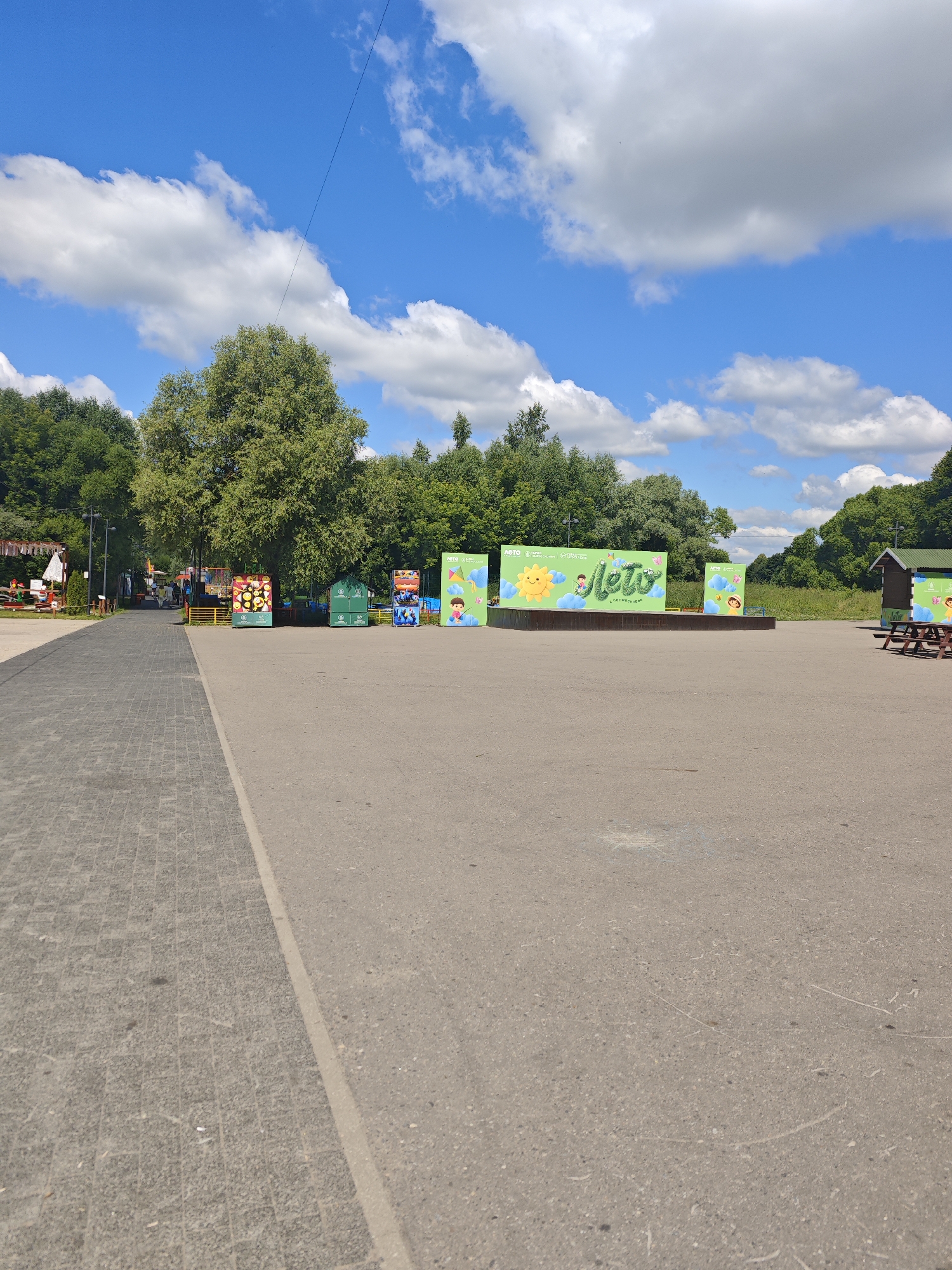 This image depicts a summer day scene in a park or outdoor recreational area, likely located in the city of Chekhov, Russia. The foreground is dominated by a paved area, with a textured gray walkway on the left and a darker asphalt surface on the right. In the mid-ground, a wooden stage is set up, decorated with large green banners featuring vibrant graphics. One banner prominently displays the word "Лето" (Summer) in a playful font, accompanied by illustrations of a smiling sun, children, and butterflies against a blue sky. To the left and right of the stage, there are several smaller, colorful stands or booths, some adorned with cartoonish imagery and logos, suggesting an event or attraction.
Behind the stage, a dense line of lush green trees forms a natural backdrop. The sky above is a bright blue, punctuated by fluffy white clouds, indicating fair weather. In the far left of the image, a section of a recreational area is visible, with more colorful structures and a glimpse of people, possibly park visitors. On the far right, a small wooden building with a green roof and picnic tables can be seen. The overall impression is of a lively, outdoor public space prepared for summer activities.