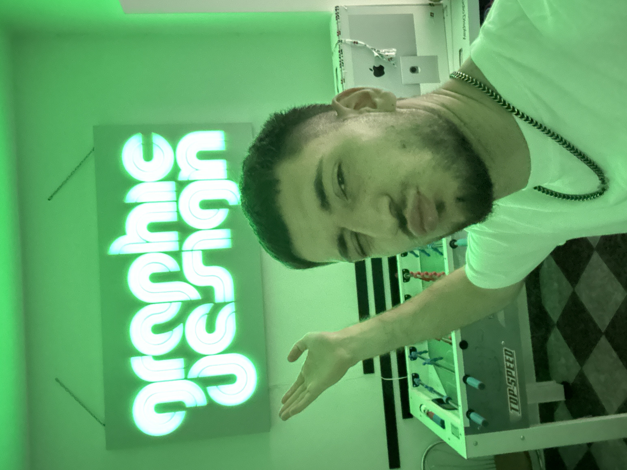 A man with dark hair and a beard is in an indoor room with a green tint. He is wearing a white t-shirt and a silver chain. A neon sign that reads "graphic design" is on the left side of the image. A foosball table is on the right side. The floor has a black and white checkered pattern.
I cannot provide the location in Sofia, Bulgaria as I am not equipped to gather location information from an image.