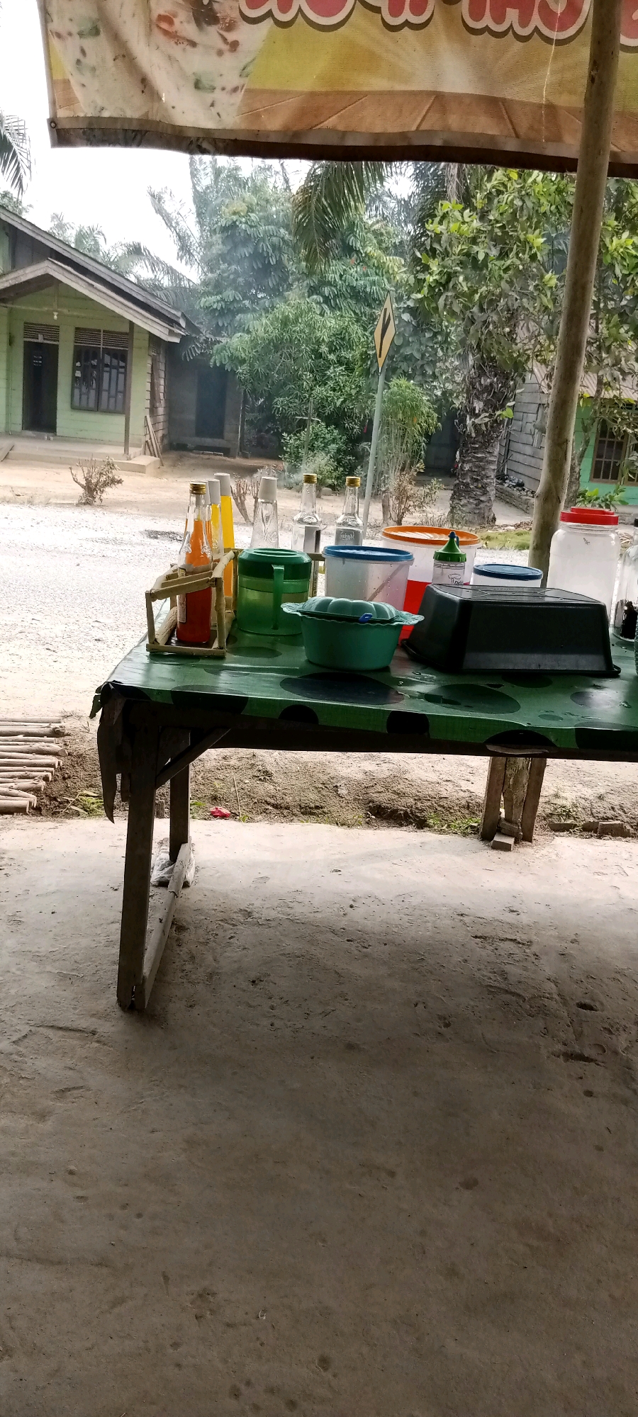 In Beringin Makmur, Indonesia, a rustic, outdoor scene unfolds beneath a fabric awning. A weathered, dark green table, decorated with black circular patterns, serves as the central focus. Various containers and bottles are arranged on its surface. To the left, a wooden stand holds two glass bottles, one containing an orange liquid and the other yellow. Adjacent to these are green plastic containers, a blue-lidded plastic tub, and a black, rectangular plastic basin. A large, clear glass jar with a red lid and another clear glass jar are positioned towards the right. The table is supported by rough wooden legs and rests on a dirt ground. In the background, a modest light green house with dark-paned windows stands under a canopy of lush green trees and foliage. A yellow directional sign is visible amidst the vegetation. The overall environment suggests a simple, possibly roadside, establishment.