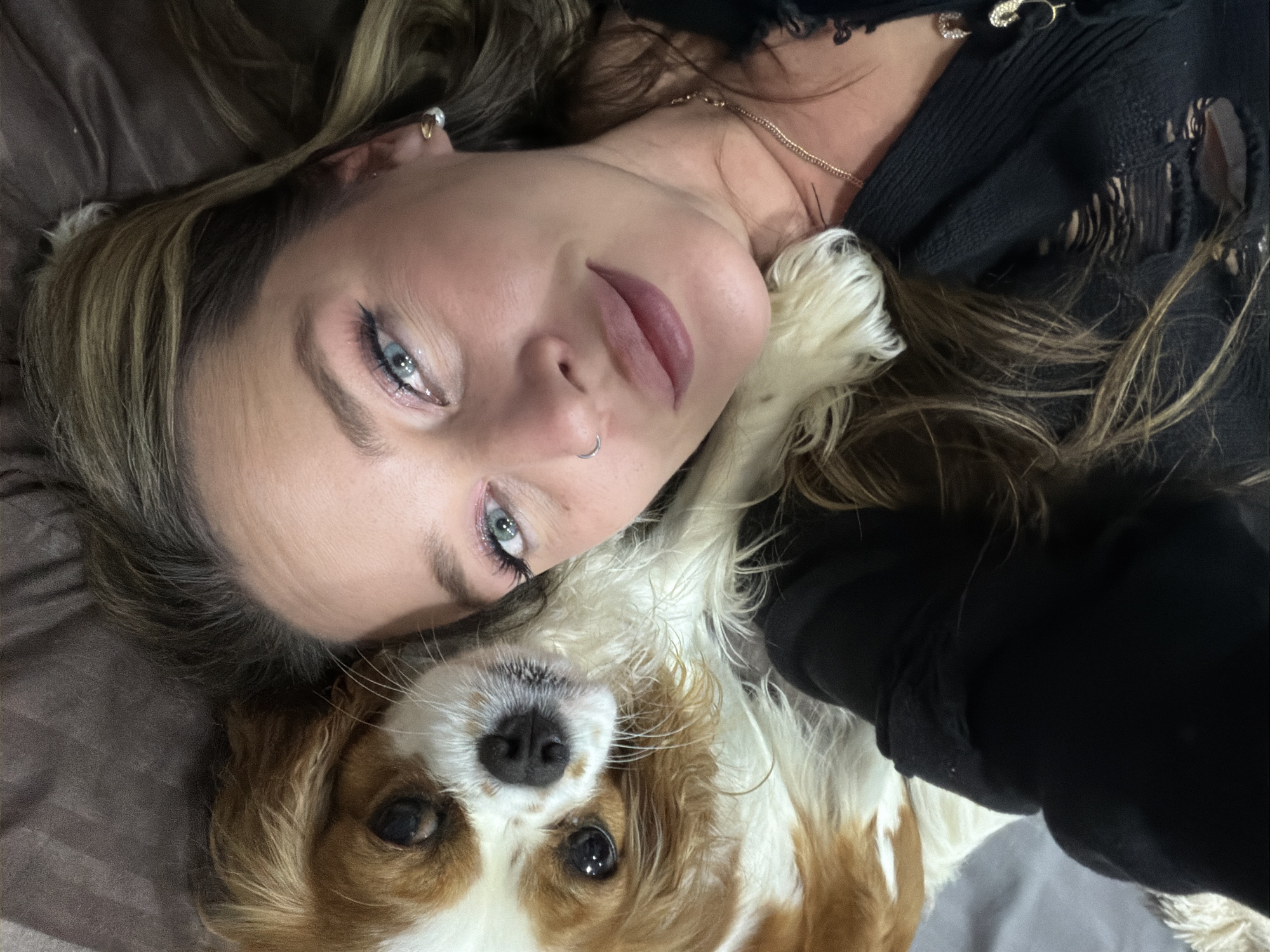 An overhead shot captures a woman and a dog lying closely together in what appears to be a relaxed, intimate setting, possibly indoors on a bed or couch, in Chaikovsky, Russia.
The woman is positioned in the upper part of the frame, looking directly up at the camera. She has long, wavy hair with light brown and blonde highlights, which spreads around her head. Her bright blue eyes are a striking feature, enhanced with dark eyeliner, mascara, and a hint of sparkle on her eyelids. She has defined eyebrows, subtle lip color, and a silver hoop nose ring. Her left ear is visible, adorned with a small gold hoop earring featuring a pearl or clear stone. She is wearing a dark, possibly black, knitted top with distressed or ripped details on the shoulder and sleeve. A delicate gold chain necklace is visible around her neck, and a decorative, jeweled safety pin is fastened to her top near her shoulder.
Lying beneath and beside her, sharing the frame, is a dog. The dog's head is visible, appearing to be a Cavalier King Charles Spaniel or a similar breed, characterized by its white and reddish-brown (blenheim) coat. Its long, floppy ears are a mix of white and brown fur. The dog has dark eyes, also looking towards the camera, and a dark nose.
The background consists of a soft, textured fabric in a muted, dark brown or grey color, contributing to the cozy and domestic feel of the scene. The overall mood is calm and affectionate, capturing a moment of companionship between the woman and her pet. No specific time of day is discernible, but the lighting appears soft and ambient. There is no visible legible text in the image.