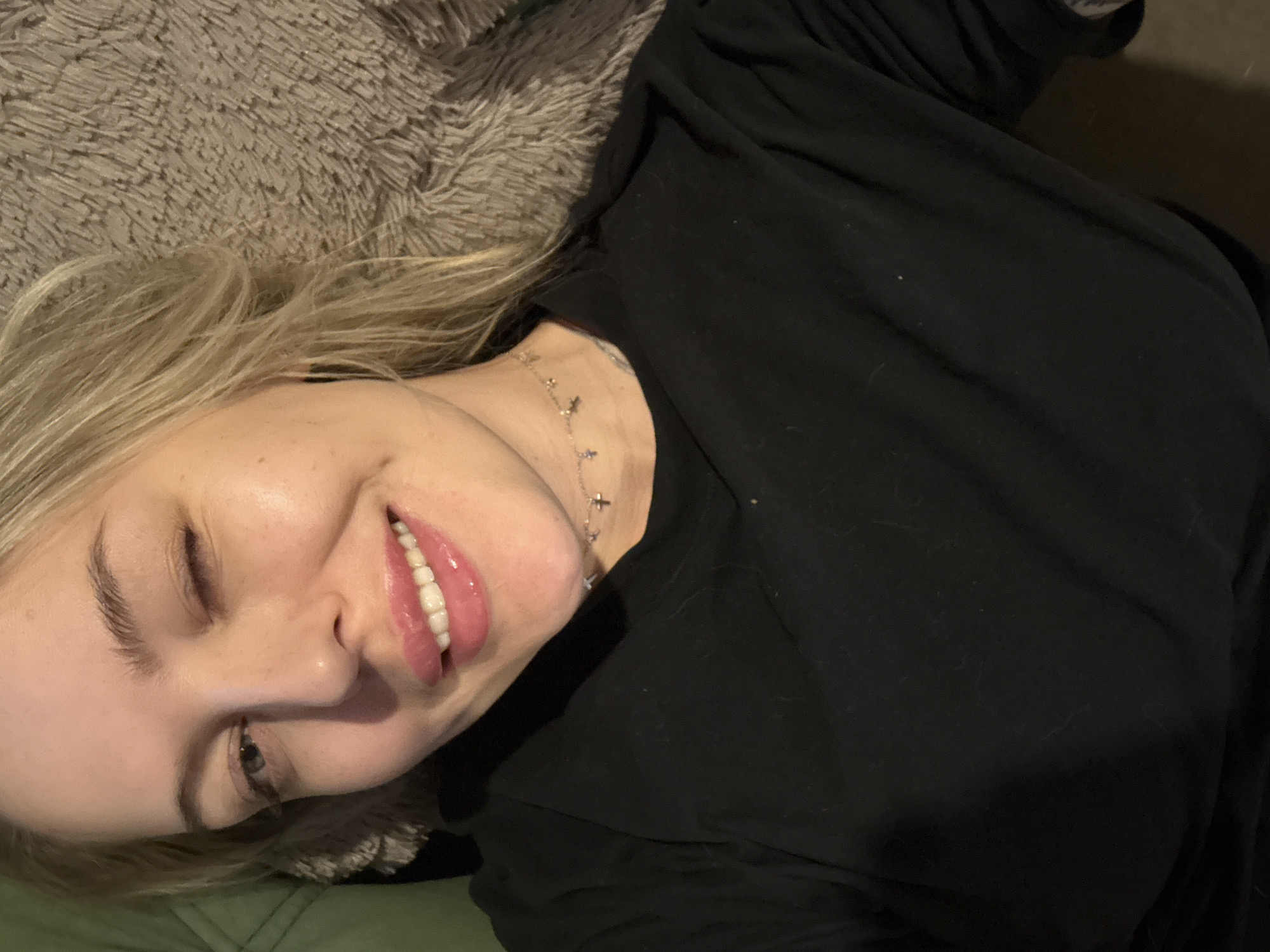 A fair-skinned woman with blond hair is lying on her side, smiling with one eye closed, suggestive of a wink. She's wearing a black long-sleeved shirt and a delicate silver necklace with cross-shaped charms. She appears to be lying on a gray, furry surface, possibly a blanket or cushion, with a hint of a green fabric visible underneath. The location in the city of Krasnoyarsk, Russia, is not discernible from the image.