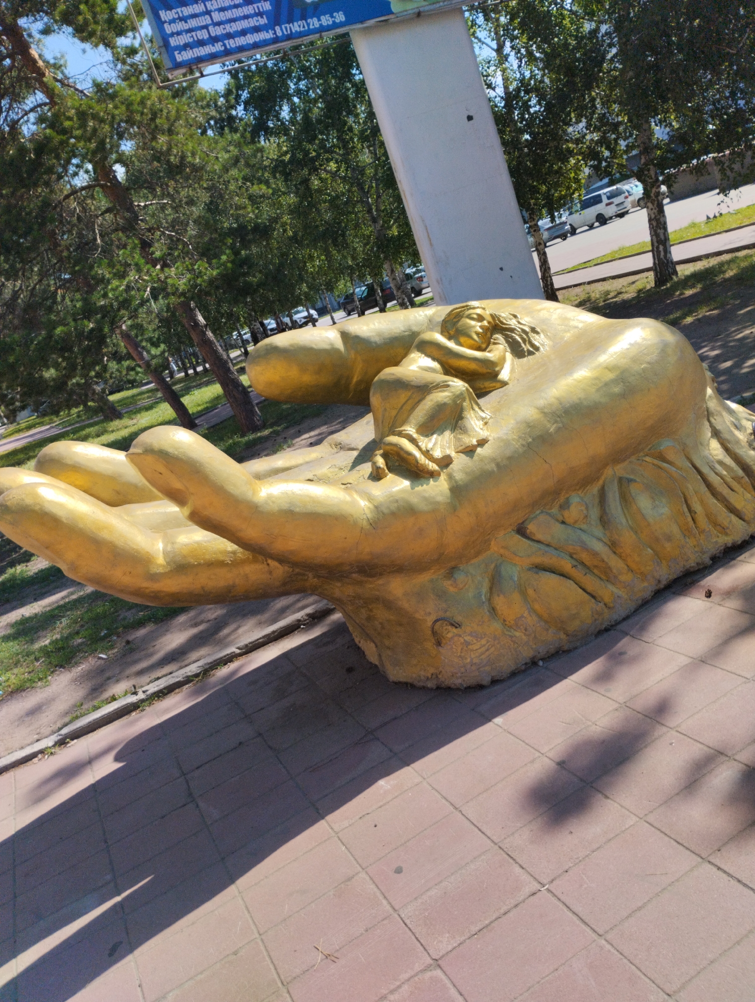 The image shows a large, golden sculpture of a hand cradling a sleeping child. The sculpture is situated outdoors, on a paved area with a brick pattern. In the background, there are trees and a road with vehicles. A sign is visible in the upper left corner with text in Cyrillic, indicating "Kostanay city" and "Regional akimat". The sculpture appears to be a public art installation.