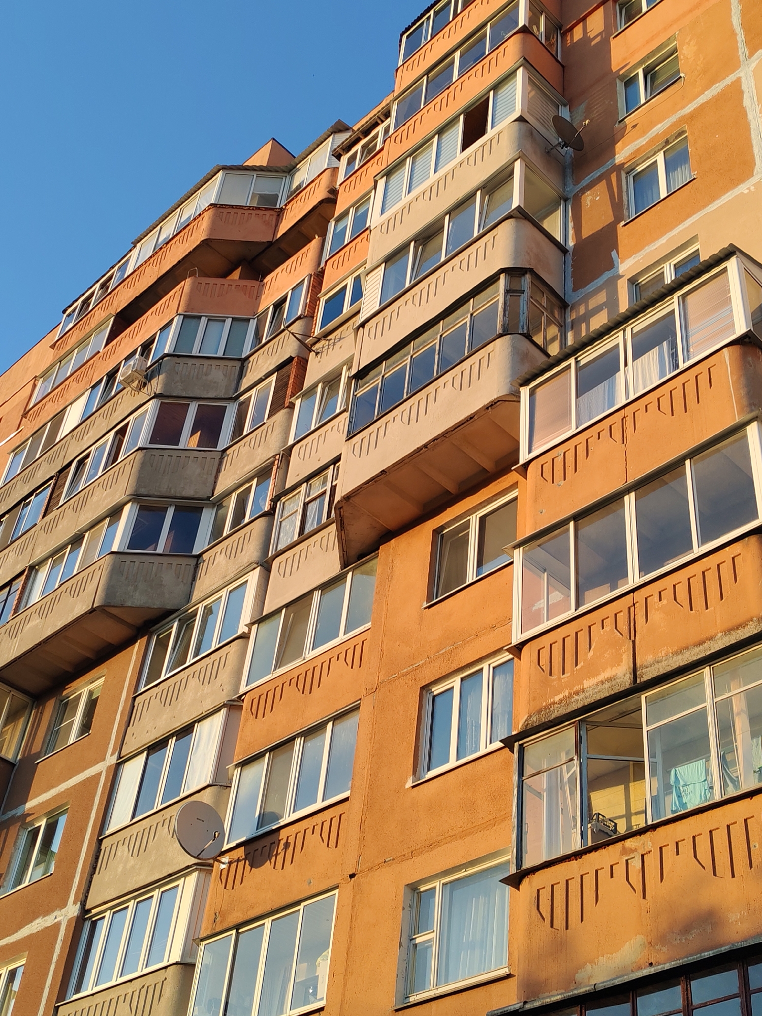The image depicts a tall, orange-painted residential building with many windows and balconies. The balconies alternate in design, some being enclosed while others are open or have a more concrete, protruding design. The building's architecture appears to be of a utilitarian style. A satellite dish is visible on one of the lower floors. The sky above is clear and blue, and the sunlight suggests it's late afternoon. The location is likely urban, with the city remaining unknown.