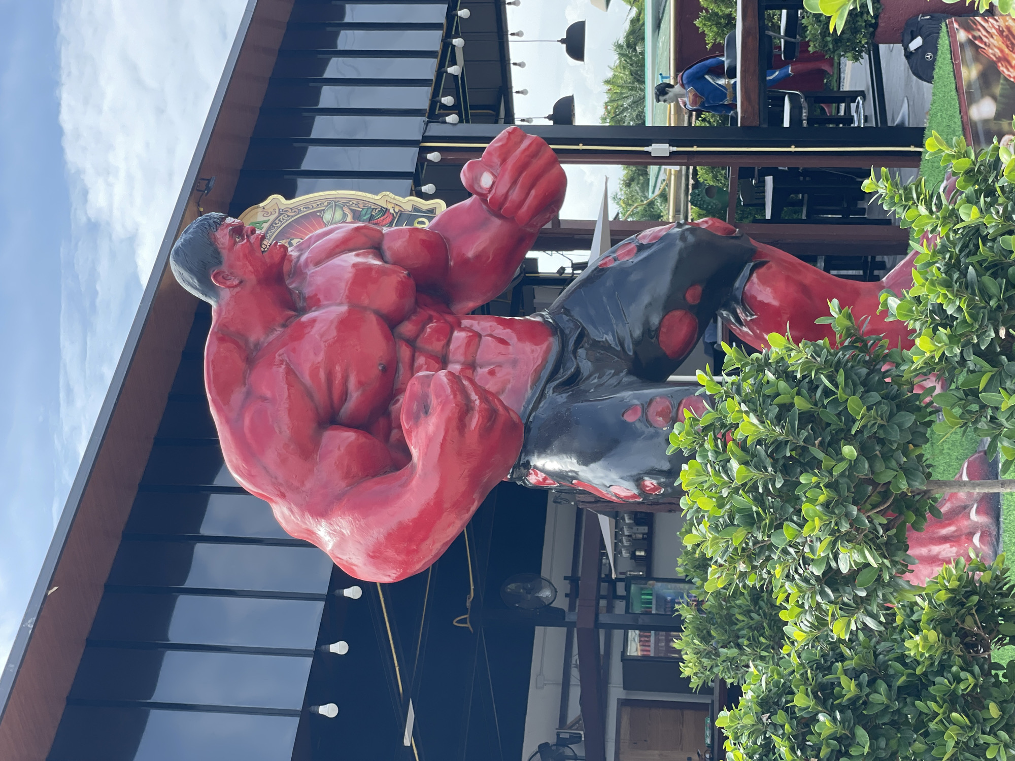 A large, red, muscular statue of the Hulk is prominently displayed outdoors in front of a modern building with a dark facade and reflective windows. The statue is wearing black shorts with red splotches. In the background, a statue of Superman can be seen standing near tables and chairs. Lush green bushes are in the foreground, partially obscuring the base of the Hulk statue. The scene appears to be located in a commercial or entertainment area, likely a restaurant or bar, given the presence of tables, chairs, and what appears to be a bar area in the background. The sky above is partly cloudy. The location is identified as Nong Prue, Thailand.