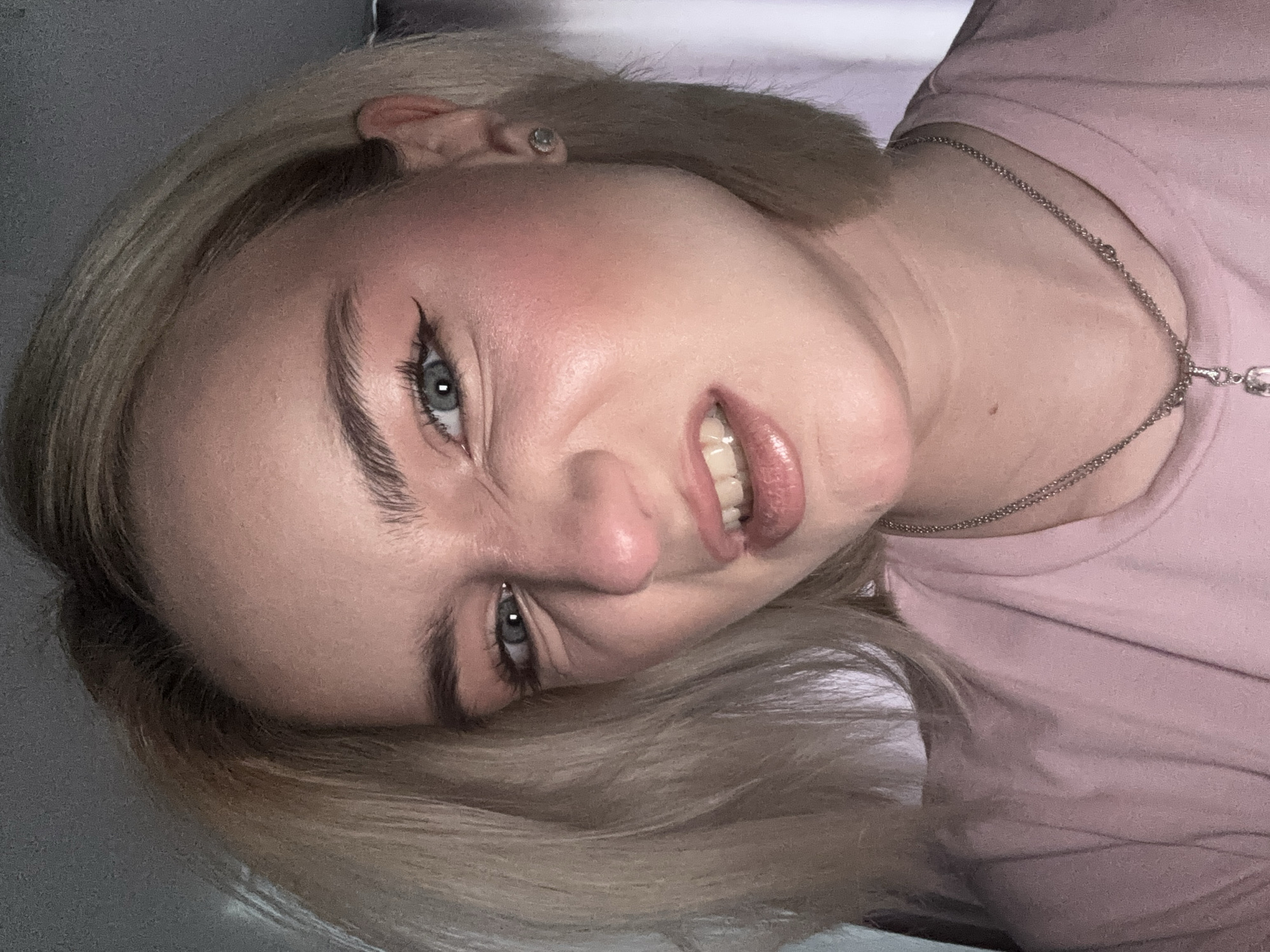 A close-up, angled portrait shows a light-skinned person with blond hair and blue eyes. The person is wearing a light pink t-shirt and a silver necklace. They are making a slightly strained facial expression. There is no information in the image to indicate a location in Vsevolozhsk, Russia, or any other specific location.