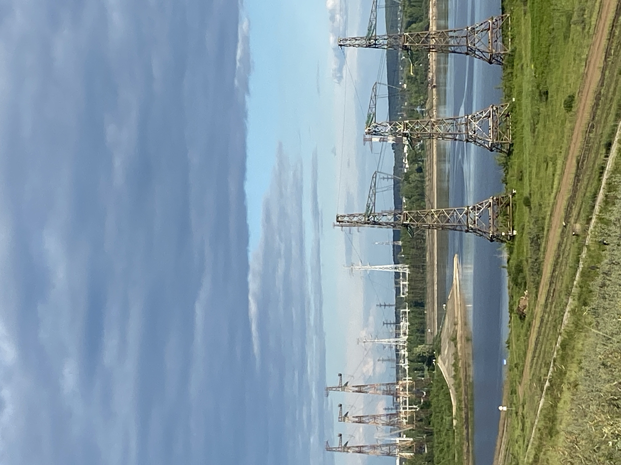 The image displays an industrial riverine landscape in Chaikovsky, Russia, under a partly cloudy sky with varying shades of blue and grey clouds. In the foreground, three large, rusty lattice-steel transmission towers stand prominently on a green grassy slope, which is traversed by a dirt track. These towers are positioned on the near bank of a wide body of water, likely a river, that dominates the mid-ground. A small, sandy peninsula or spit of land extends into the water on the left side of the frame. The far bank of the river is lined with dense green trees and vegetation. In the background, an extensive array of numerous additional power transmission towers of various designs (including both white and darker lattice structures) stretches across the horizon, indicating a significant electrical power generation or distribution facility. These distant towers are set against a backdrop of distant tree-covered land. No people are visible in the scene.
