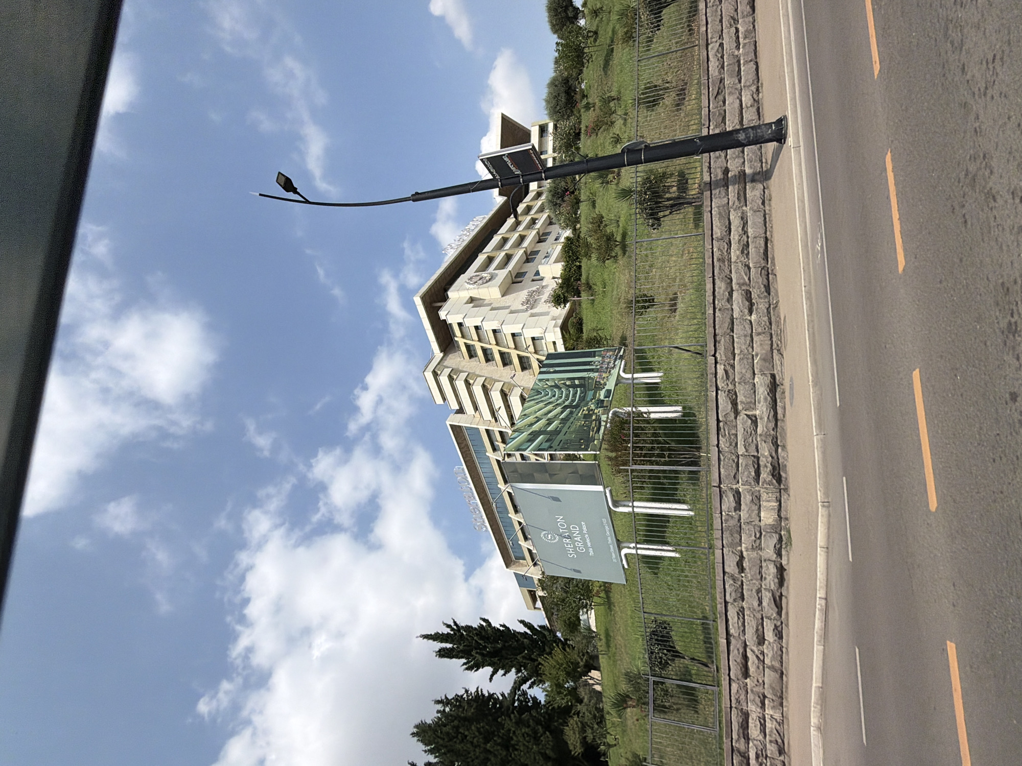 The image captures a frontal view of the Sheraton Grand Tbilisi Metechi Palace, a large hotel situated on a grassy hillside in Tbilisi, Georgia. The hotel's distinctive architecture, with its tiered levels and angular facade, is prominent against a bright blue sky with scattered white clouds. A large billboard advertising the hotel is positioned in front of the building, displaying the Sheraton Grand logo and an image of the hotel's interior. Adjacent to the billboard is a tall, dark lamppost with a small rectangular sign attached to it.
The foreground of the image shows a paved road with yellow lane markings. A low stone wall borders the road, with a metal fence above it separating the road from the hotel's grounds. Lush green grass and various trees and bushes cover the hillside leading up to the hotel. The overall scene depicts a sunny day, with clear visibility of the hotel and its surroundings.