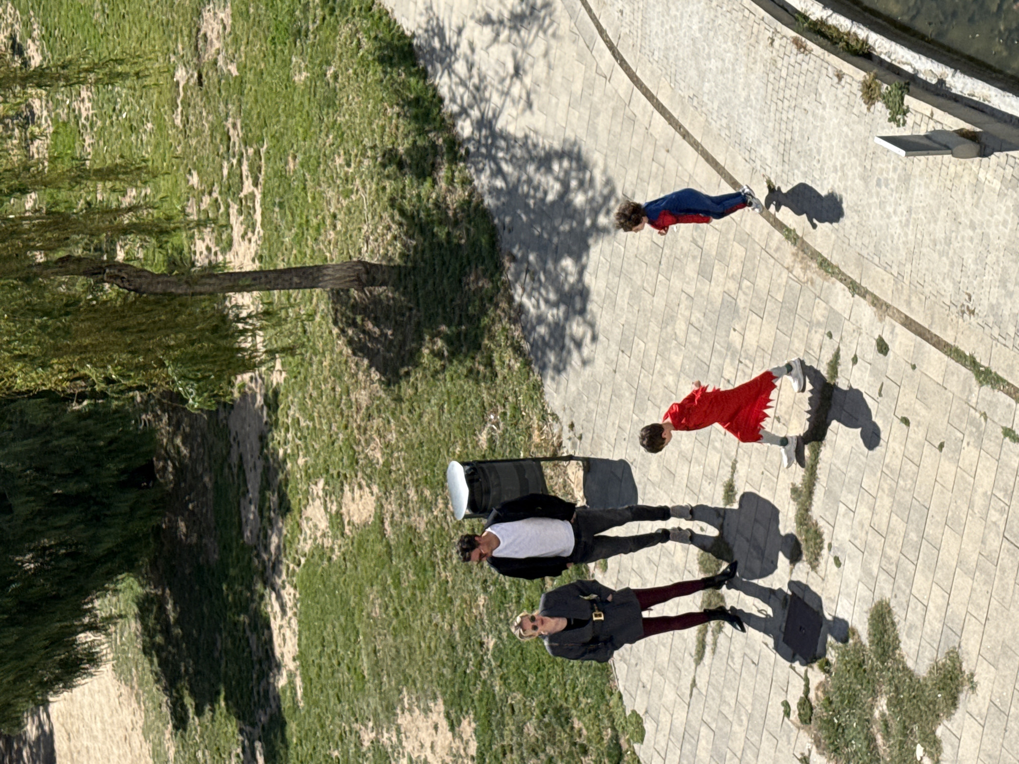 The image, taken from above, shows a group of four people standing on a paved walkway adjacent to a grassy area with a tree. The location is likely Barcelona, Spain, based on architectural features. The walkway runs alongside what appears to be a river or canal, with a concrete edge. The people are oriented in a line. One person is wearing a blue and red suit. Another individual wears a red dress, and a third is dressed in a white shirt and dark trousers. The fourth person is wearing a grey dress. Their shadows are cast on the paved surface. One person is next to a black suitcase.