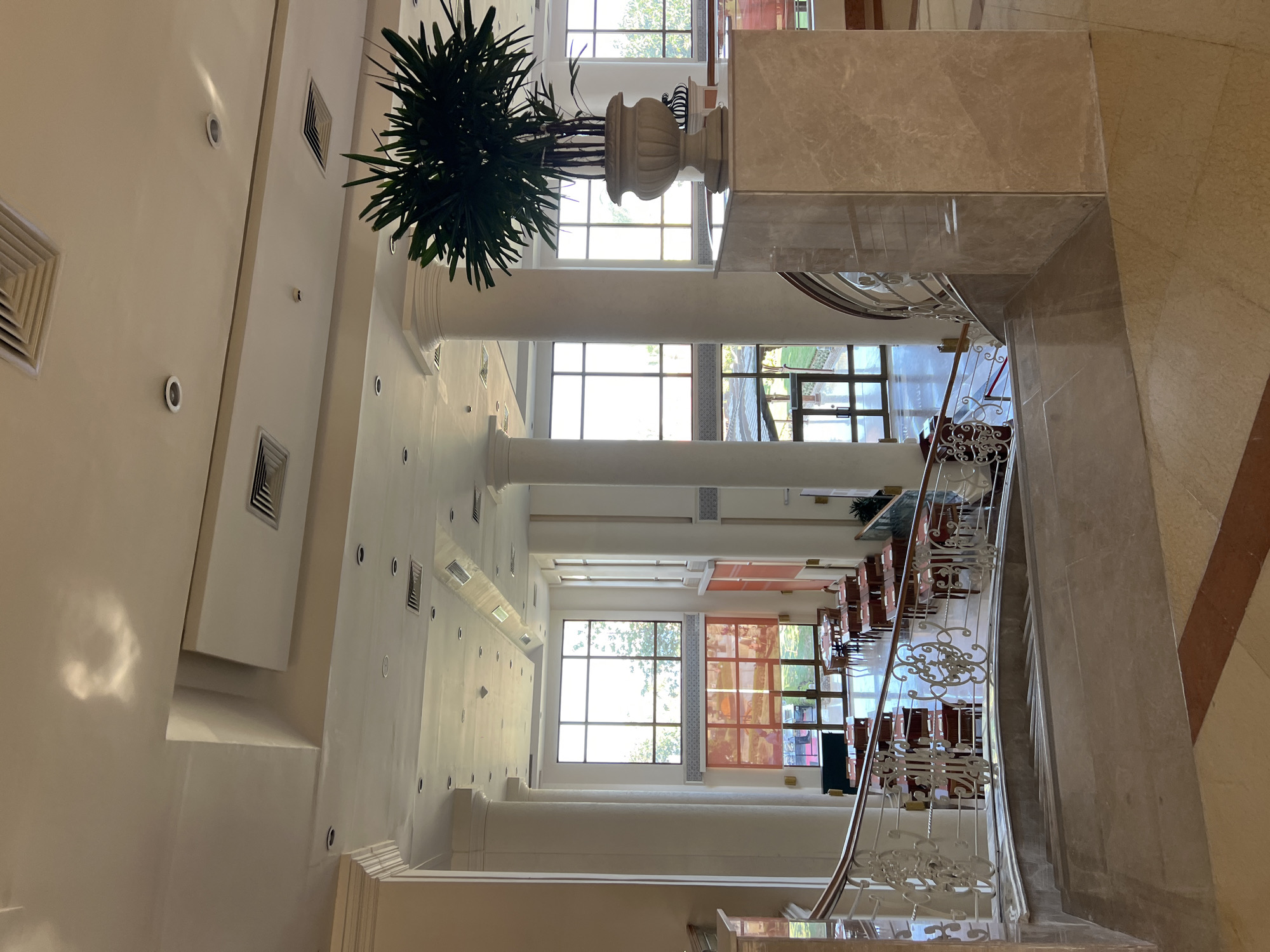 The image depicts a grand hotel lobby in Sharm El Sheikh, Egypt. The scene is dominated by a large, open space with high ceilings and numerous tall, white columns. A sweeping staircase with ornate white railings curves into the foreground, leading down from what appears to be an upper level. The staircase is lined with marble.
Through large windows in the background, a bright, sunny day is visible, with trees and greenery suggesting an outdoor area beyond. The lobby itself has a polished marble floor with geometric patterns. Several dining tables and chairs are arranged in the lower level of the lobby, indicating a restaurant or dining area. A large potted plant with dark green foliage sits on a marble pedestal to the right of the frame, adding a touch of natural decoration. The lighting appears to be a mix of natural light from the windows and recessed ceiling lights. The overall impression is one of spaciousness, elegance, and a well-maintained hospitality environment.