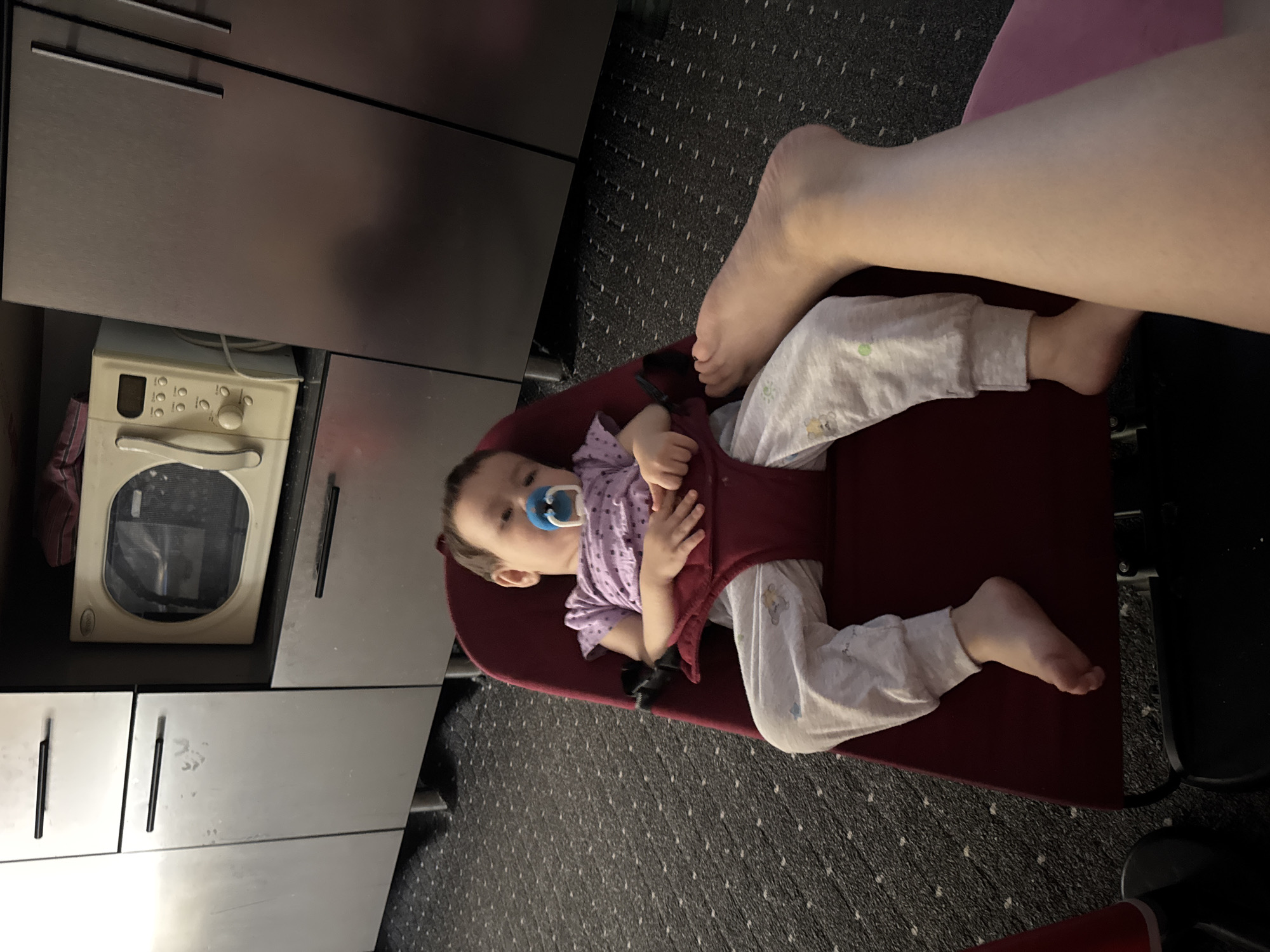 A toddler is sitting in a red bouncer chair with a blue pacifier in their mouth. The toddler is wearing a purple t-shirt and grey sweatpants with cartoon characters. The child's feet are visible. In the background, there are kitchen cabinets and a microwave. A person's leg is visible in the foreground, suggesting the photo was taken from their perspective. The scene appears to be indoors, likely a kitchen or living area. The carpeted floor is dark with small white dots. The location is not specified within the image or its metadata.