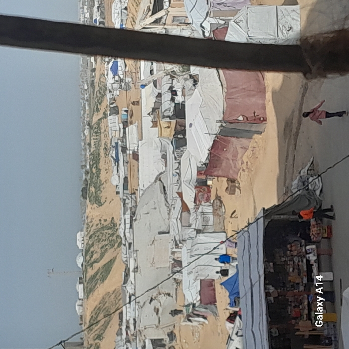 An elevated view captures a densely populated, informal settlement in Khan Yunis, Palestinian Territory. The scene is dominated by hundreds of makeshift shelters constructed from white tarpaulins, plastic sheeting, and fabric stretched over frames, sprawling across undulating sandy terrain.
In the immediate foreground, a dark vertical beam partially obstructs the right side of the frame. To the lower left, a makeshift market stall, covered by a dark tarp, displays various goods on tables. Near this stall, one individual in an orange top and dark pants stands, facing right. Another person, a child in a pink top and dark pants, walks away from the stall down a dirt path towards the right. A dark power line or cable traverses the mid-foreground diagonally.
The midground features numerous closely packed shelters on sandy ground, with visible dirt paths winding between them. Several individuals are present: three figures are visible in the center-left, one in a blue top, standing or walking near a blue tarp-covered area. Indistinct human figures are scattered throughout the camp.
In the background, sandy hills rise, exhibiting sparse patches of green vegetation. More shelters extend across and beyond these hills, indicating the expansive nature of the camp. A tall, light-colored tower, possibly a communication mast, stands atop one of the hills on the upper left. The sky above is a light, hazy grey-blue. The overall environment is arid and dusty.