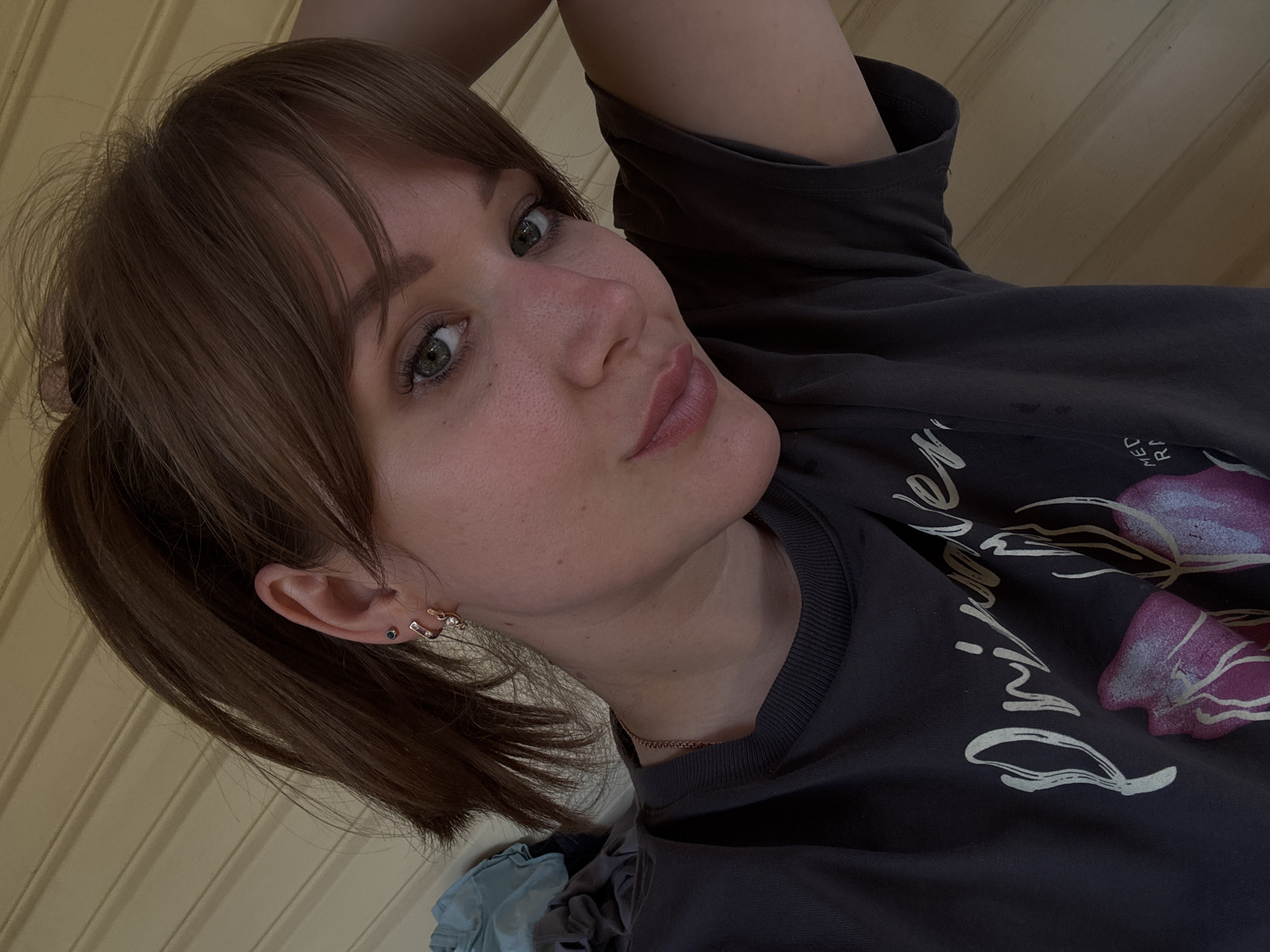 A female individual, approximately 20-30 years old, with medium brown hair styled in a high ponytail and bangs, looks directly at the camera with a gentle smile. Her complexion is fair with visible freckles across the nose and cheeks, and her eyes are green, enhanced by dark lashes and subtle eyeliner. Her lips are a muted rose color. A thin, gold-colored chain necklace rests around her neck. The visible left ear displays an upper lobe piercing with a small dark stud and a lower lobe adorned with two rose gold-toned hoop earrings; the larger hoop features small clear stones, and a smaller plain hoop is positioned beneath it. She wears a dark grey, short-sleeved crewneck t-shirt. The shirt's front features a graphic design with light-colored script partially spelling "Pri... Ne..." above a prominent magenta and purple abstract floral motif, with "MED RA" visible below. Her left arm is raised, with the hand positioned behind her head, out of frame. A pile of dark and blue textile materials is partially visible to the left. The background consists of horizontally oriented light beige wooden paneling. The scene is illuminated by soft, ambient light. The location is Chaikovsky, Russia.
