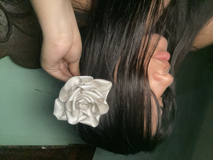 A woman's profile with dark hair, positioned upside down, is seen against a muted green background. Her lips are slightly parted. A large, artificial white rose is placed near her ear, seemingly held by her hand, which is positioned above the flower. The backdrop features textured elements, suggesting a surface or object.