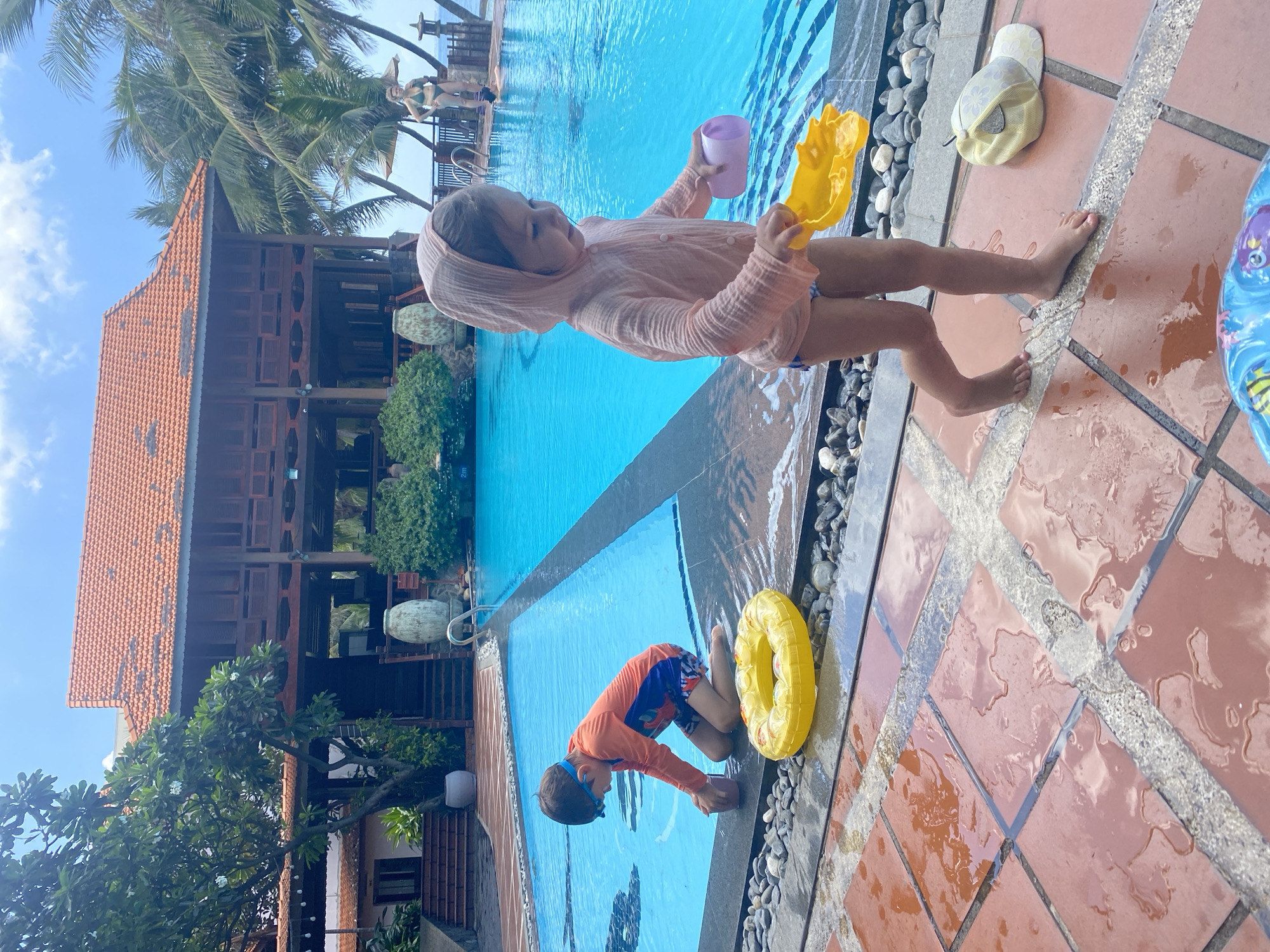 This image captures an outdoor swimming pool area in Phu Thuy Ward, Vietnam, on a bright, partly cloudy day. In the foreground, two children are observed near the pool's edge. A boy, estimated to be 5-8 years old, wearing an orange long-sleeved rash guard, blue patterned swim shorts, and blue swimming goggles, kneels in the shallow end of the pool, interacting with a purple cup. Adjacent to him, a younger girl, estimated to be 2-4 years old, stands barefoot on wet, reddish-brown square tiles. She wears a light pink hooded top over patterned swim bottoms and holds a yellow plastic shovel in her right hand and a purple plastic cup in her left. A deflated yellow inflatable ring rests on the wet tiles near the boy, and a yellow baseball cap with white floral patterns lies near the girl's feet. A border of grey and white pebbles separates the tiled ground from the pool's edge. The bright blue swimming pool extends into the mid-ground, featuring an infinity edge towards the right. In the background, a multi-story building with a prominent reddish-orange tiled roof and dark wooden architecture is visible, surrounded by abundant green foliage, including palm trees. Two large, light-colored ornate vases are positioned near the building and the pool. Further right in the background, a woman wearing a dark green two-piece swimsuit stands by the infinity pool, looking towards the left. Beyond the pool and trees, a clear blue sky meets the horizon of a body of water, consistent with an ocean view.