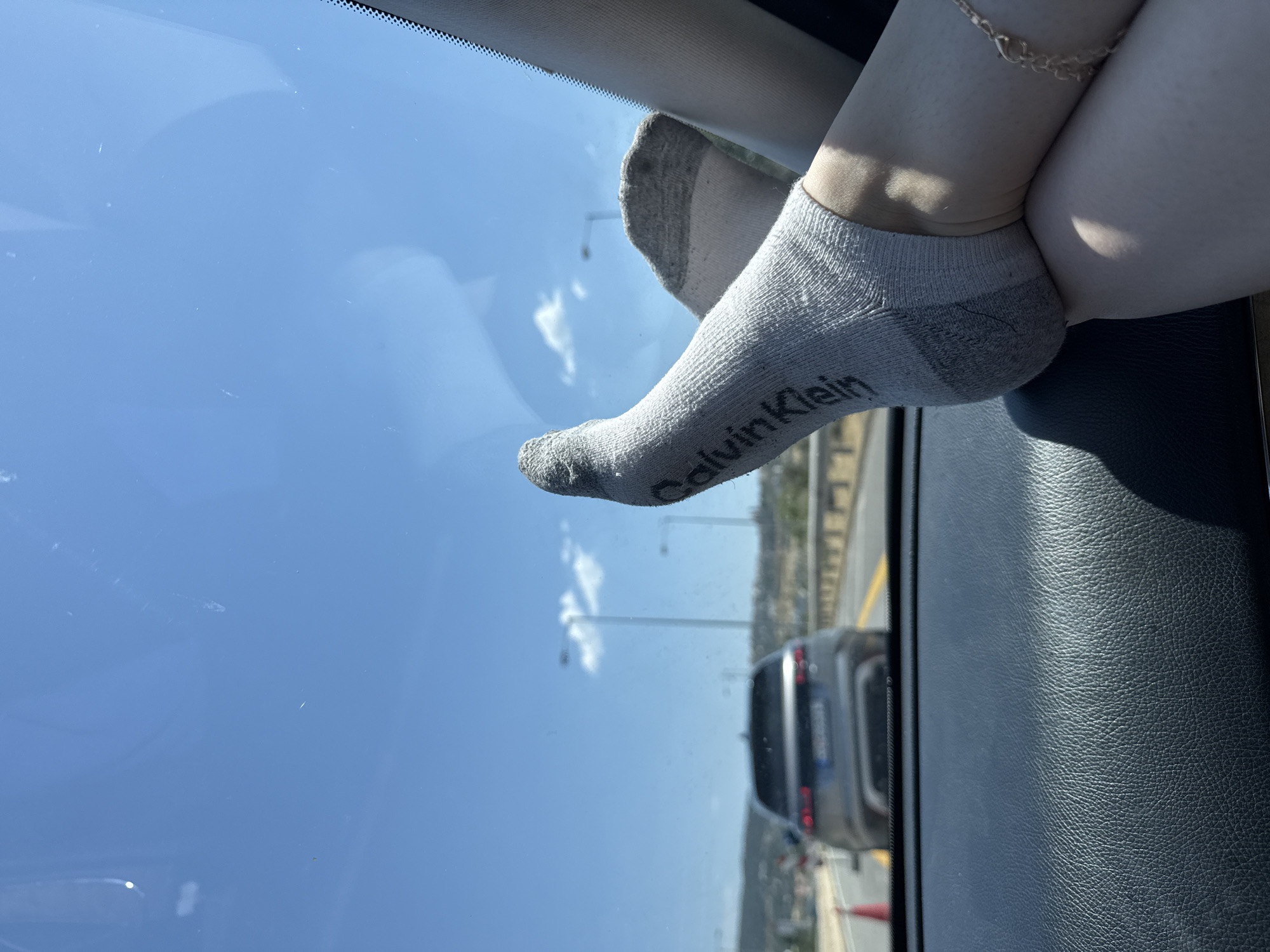 A person's feet, wearing gray Calvin Klein ankle socks, are resting on the dashboard of a car. One foot is bent, with the heel of the sock clearly visible, while the other is extended, showing the sock's toe. A delicate gold anklet is worn on the ankle of the extended foot.
The car appears to be on a highway, as evidenced by the road markings and other vehicles visible through the windshield. The sky is a bright blue with scattered white clouds, suggesting it is daytime with clear weather. The sunlight casts shadows on the dashboard and the person's legs.
The background shows a glimpse of a landscape with hills and a bridge, consistent with a highway setting. The overall impression is a casual moment during a car journey, likely in Greece given the location context. The scene is captured from a low angle, focusing on the feet and the view outside.