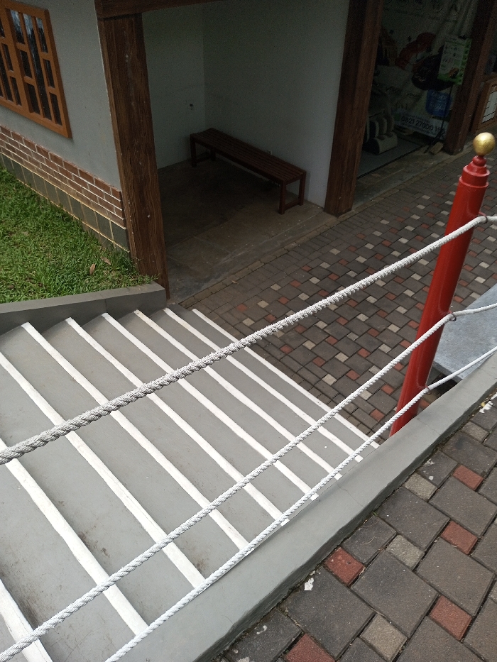 The image depicts an outdoor architectural scene in Tân Kỳ Commune, Vietnam, captured from an elevated perspective looking down. The primary subject is a descending gray concrete staircase with white painted lines on each tread. A light gray rope railing system, composed of three parallel strands, runs diagonally alongside the stairs, attaching to a prominent red cylindrical post topped with a gold spherical finial on the right. This post stands on a paved surface.
To the left of the staircase, a section of a building is visible, featuring a light gray wall above a brown brick base course. A brown wooden window frame with horizontal slats is set into this wall. Below an overhang of the building, a recessed alcove contains a dark brown wooden slatted bench against a light gray back wall. The floor of this alcove is concrete.
The ground around the building includes green grass to the left of the brickwork and a patterned walkway of interlocking concrete pavers in shades of gray and reddish-brown, situated at the top of the stairs and extending to the right. Beyond the recessed alcove, a dimly lit area is visible through an opening, revealing indistinct objects or equipment. No people or specific actions are depicted in the scene.