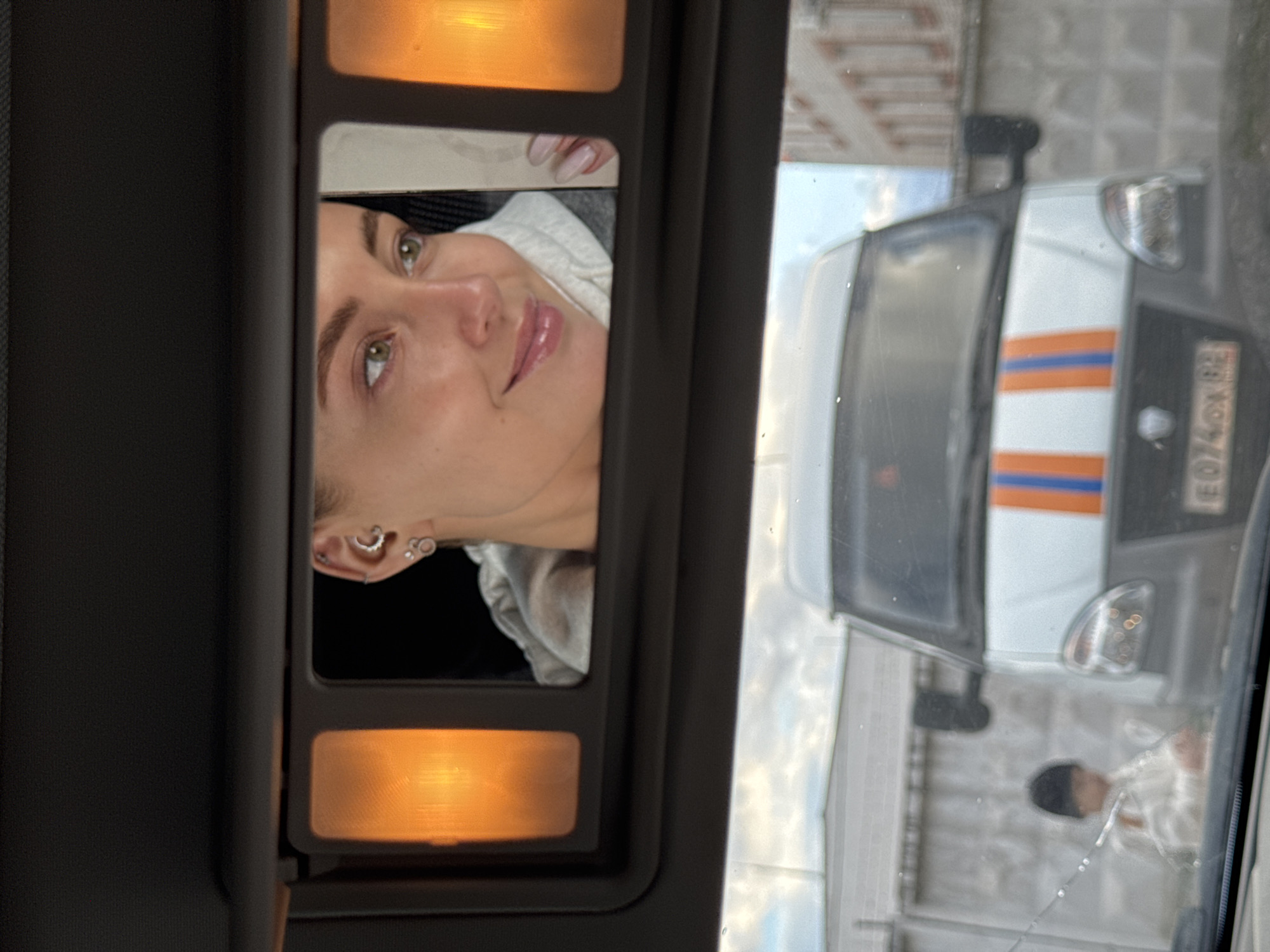 The image, taken from inside a vehicle in Chaikovsky, Russia, presents a dual reflection. In the foreground, a lowered sun visor reveals a vanity mirror reflecting the upper half of a woman's face. She has light skin, green/hazel eyes, and light brown hair. Her lips are adorned with a light pink gloss, and multiple ear piercings, including a heart-shaped stud, are visible. Her fingers, with light-colored manicured nails, are near her face. She appears to be wearing a light-colored hooded top. Two warm, amber-toned lights illuminate the mirror from above and below.
Simultaneously, the car's front windshield reflects the exterior scene. A white vehicle with prominent orange and blue horizontal stripes across its hood and front is clearly visible, suggesting it is a service or emergency vehicle. Its license plate is partially readable as "E074OX 82," indicating a Russian registration with "82" as the region code. A faint, blurred reflection of a person wearing a dark cap is also visible in the lower part of the windshield. The external background, as seen through the reflection, shows a cloudy daytime sky above what appears to be a concrete or paved structure, possibly an urban or industrial setting. The overall impression is a moment of self-reflection and observation from within a stationary vehicle during the day.