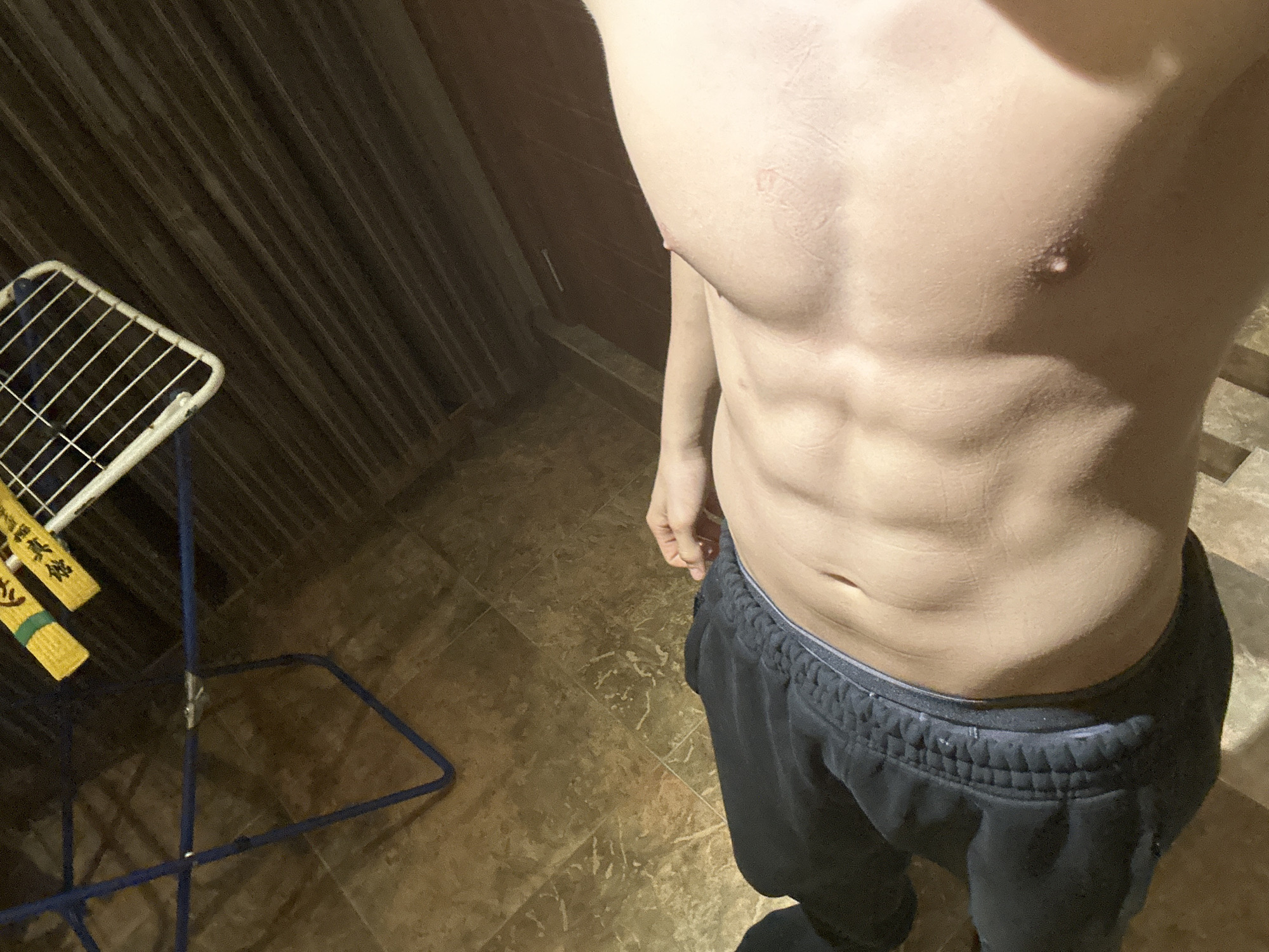 A person with a visible torso and abdominal muscles is standing in what appears to be an indoor space. They are wearing dark-colored sweatpants. To the left of the person, there is a blue and white clothes-drying rack. The flooring is tiled in a light brown marble-like pattern. The background includes what looks like dark vertical siding.
There is no evidence in the image to identify the location as Burgas, Bulgaria.