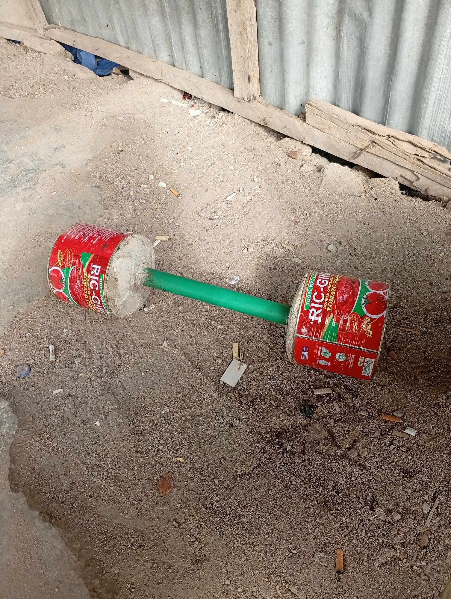 The image depicts a makeshift dumbbell lying on a dirt surface. The dumbbell is constructed from two red cans labeled "RIC-GIK DOUBLE CONCENTRATED TOMATO PASTE", connected by a green pipe. The cans appear to be filled with a gray substance, possibly cement or plaster. The surrounding dirt is littered with debris, including cigarette butts, small pieces of paper, and other unidentifiable objects. In the background, there is a corrugated metal wall, with wooden supports and debris. The scene appears to be outdoors, potentially in a neglected or impoverished area. There is no visual confirmation of the location of Adankolo, Nigeria.