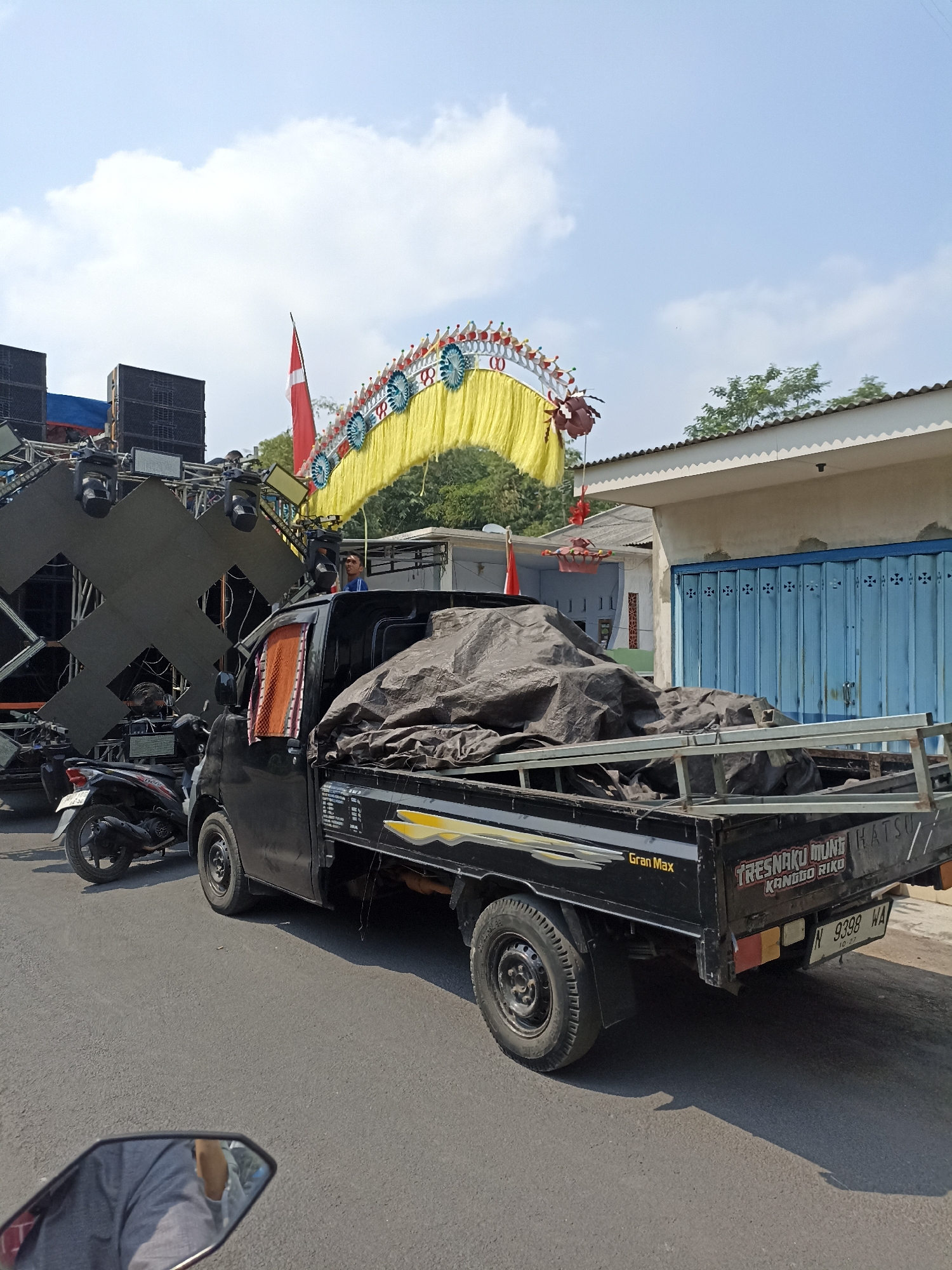 In Karangbendo, Indonesia, a black pickup truck is parked on an asphalt road. The truck's bed is covered with a large dark tarp. To the left of the truck, a black motorcycle is parked. In the background, a large sound system with speakers and lights is visible, along with a yellow decorative arch adorned with red and blue ornaments. A red flag is attached to a pole near the arch. Behind the sound system and arch, there is a building with a blue roll-up door and a white facade. A man is standing on a platform behind the truck, looking towards the left. The sky is blue with scattered clouds.