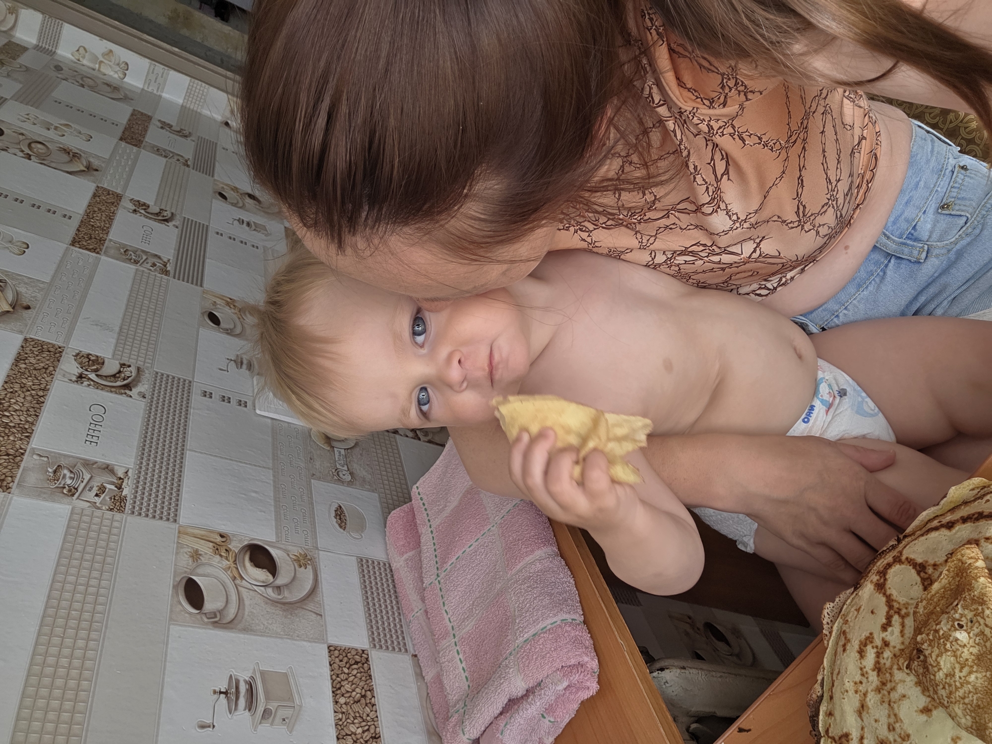 A woman is embracing a baby, leaning in to kiss the baby's forehead. The baby, held in the woman's arms, looks towards the camera with a piece of food in its hand. The baby is wearing a diaper. In the foreground, a stack of pancakes is visible. The background features a tiled wall with a coffee-themed design, including images of coffee cups, coffee beans, and coffee grinders, and the word "COFFEE" is visible on one of the tiles. The overall scene appears to be indoors, possibly a kitchen, given the tiles and the presence of food. The lighting suggests it might be daytime. The woman has long brown hair and is wearing a light-colored, patterned top and denim shorts. The baby has light hair and blue eyes.