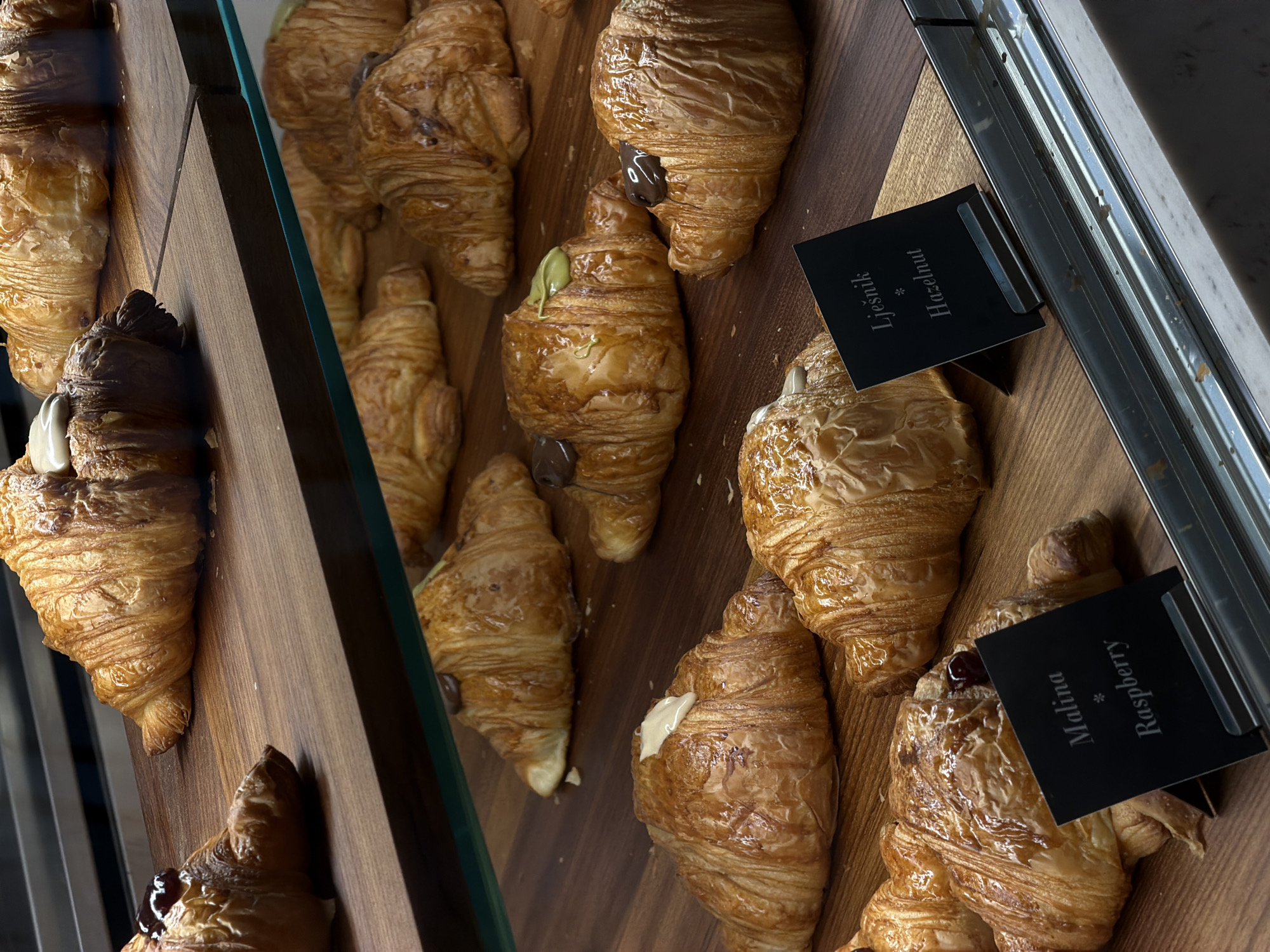 The image shows a bakery display in Budva, Montenegro. A selection of croissants with various fillings is arranged on wooden shelves behind a glass partition. From left to right, the croissants appear to be filled with almond, chocolate, pistachio, hazelnut, vanilla, and raspberry. Small, black labels identify the "Hazelnut" and "Raspberry" varieties in both Croatian ("Ljesnik", "Malina") and English. The bakery case has a metal frame. The background includes the bakery's interior.