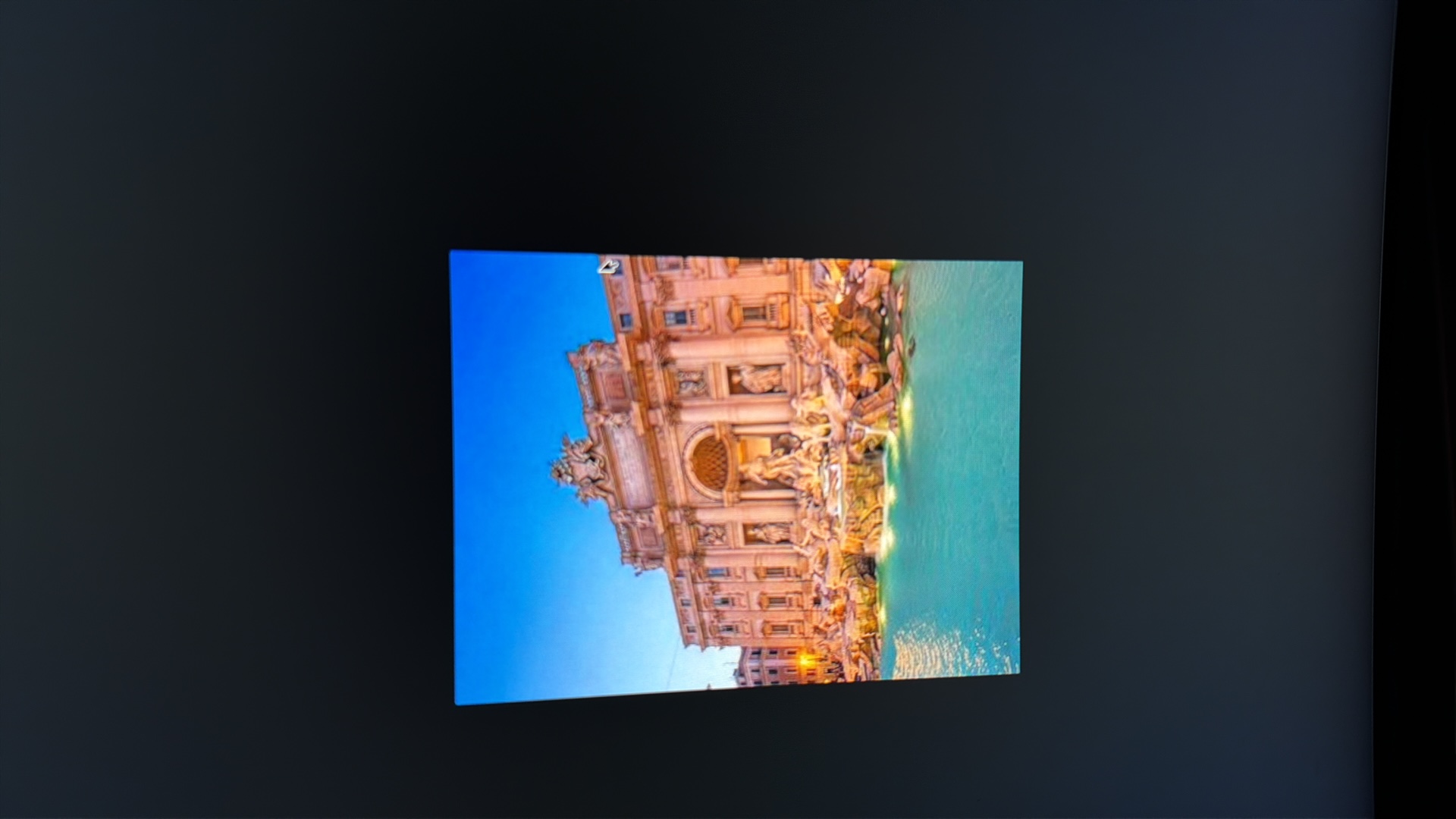 The image displays a nighttime view of the Trevi Fountain in Rome, Italy. The fountain is illuminated and features statues, intricate carvings, and cascading water into a large pool. The architecture of the surrounding buildings is visible behind the fountain. The sky is a deep blue, suggesting it is dusk or early night. The foreground is dominated by the turquoise water of the fountain. There are no people visible in the image. The photograph appears to be taken from a slightly elevated perspective. There is no indication that this image is located in Kafr Yasif, Israel.